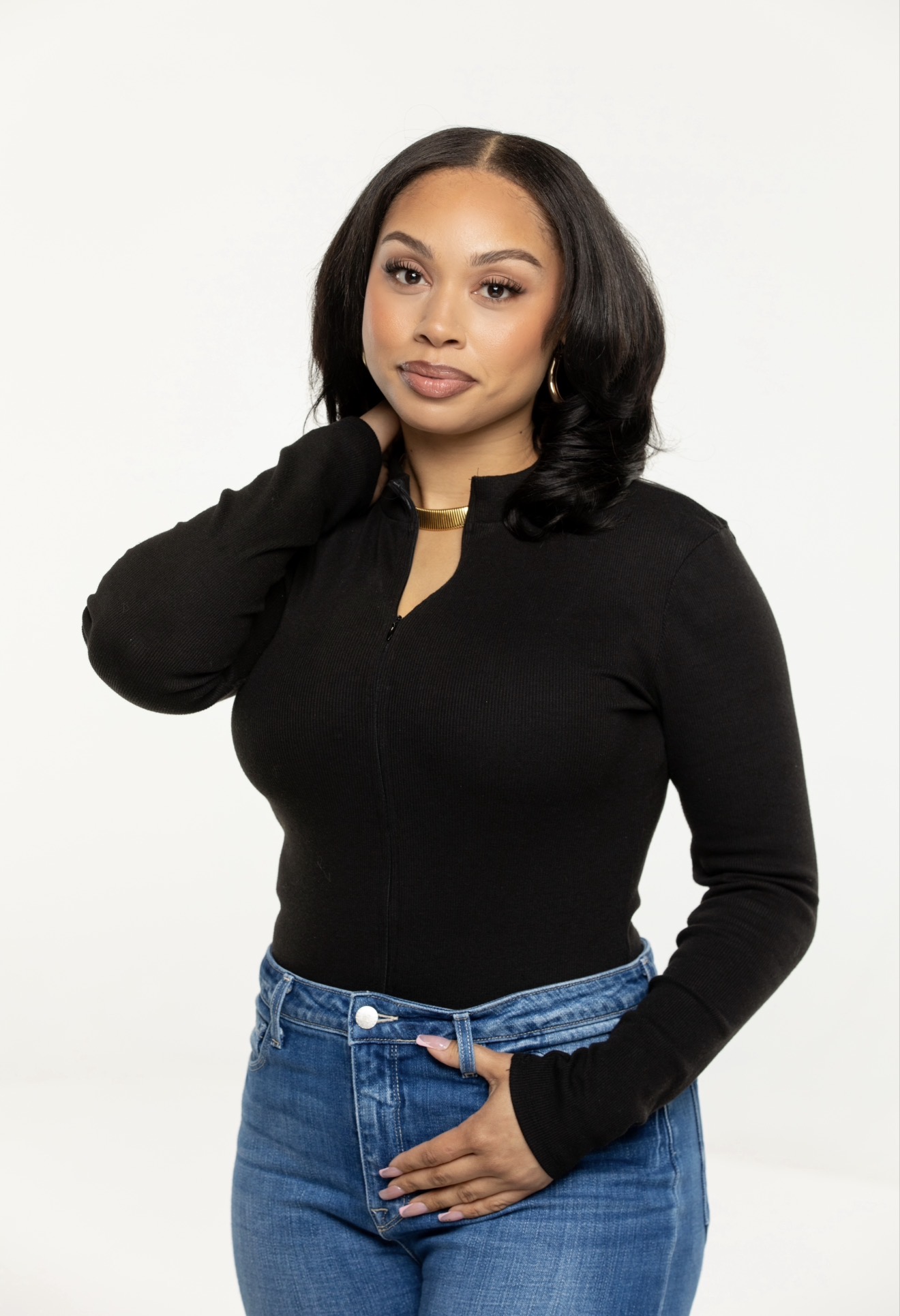 Woman with shoulder-length black hair wearing a black top and blue jeans, posing with one hand near her neck.
