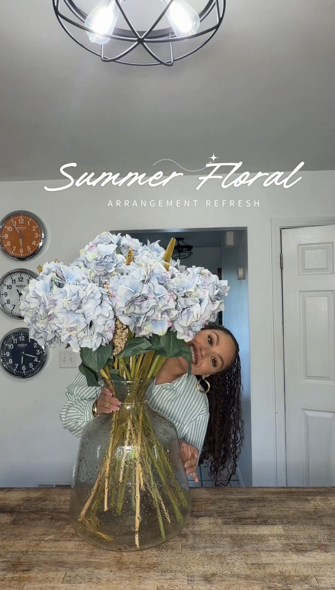 Person holding a large bouquet of white and purple flowers in a glass vase, standing in a room with clocks on the wall.