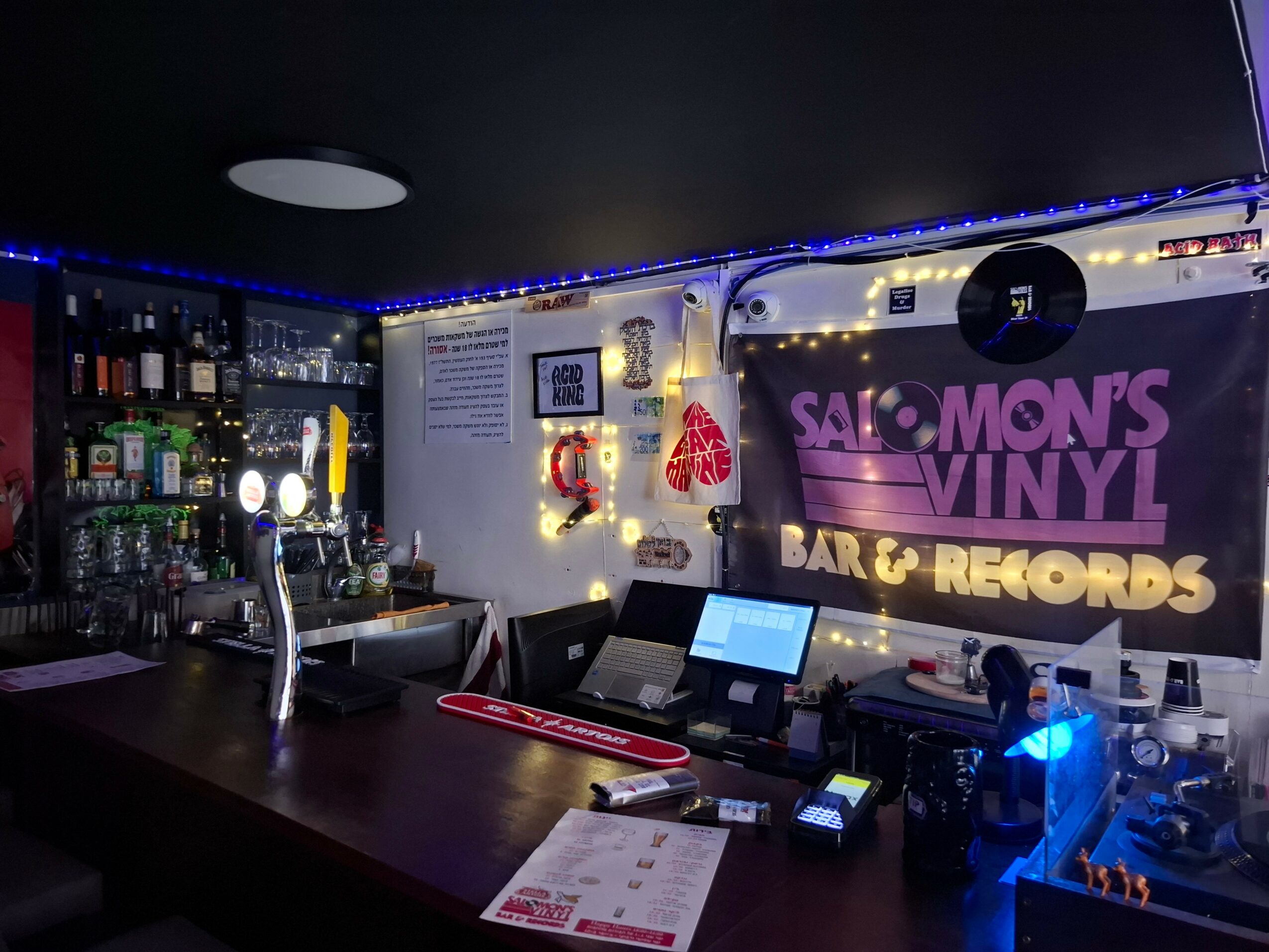 Bar with illuminated signs, shelves of bottles, and a neon sign reading 'Salmon's Vinyl Bar & Records'.