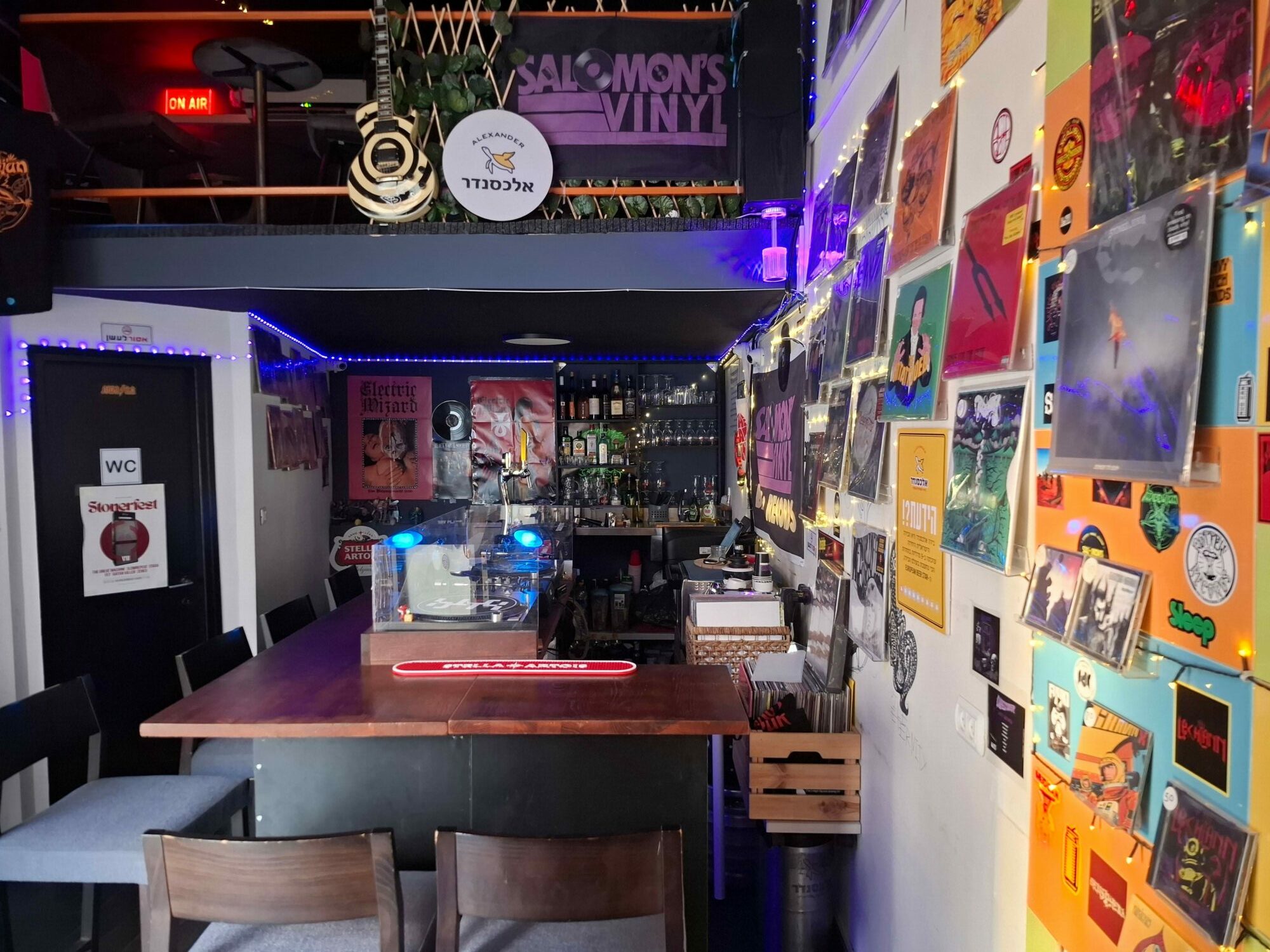 Indoor space with tables, chairs, and colorful wall decorations, including posters and photos, with a bar area in the background.