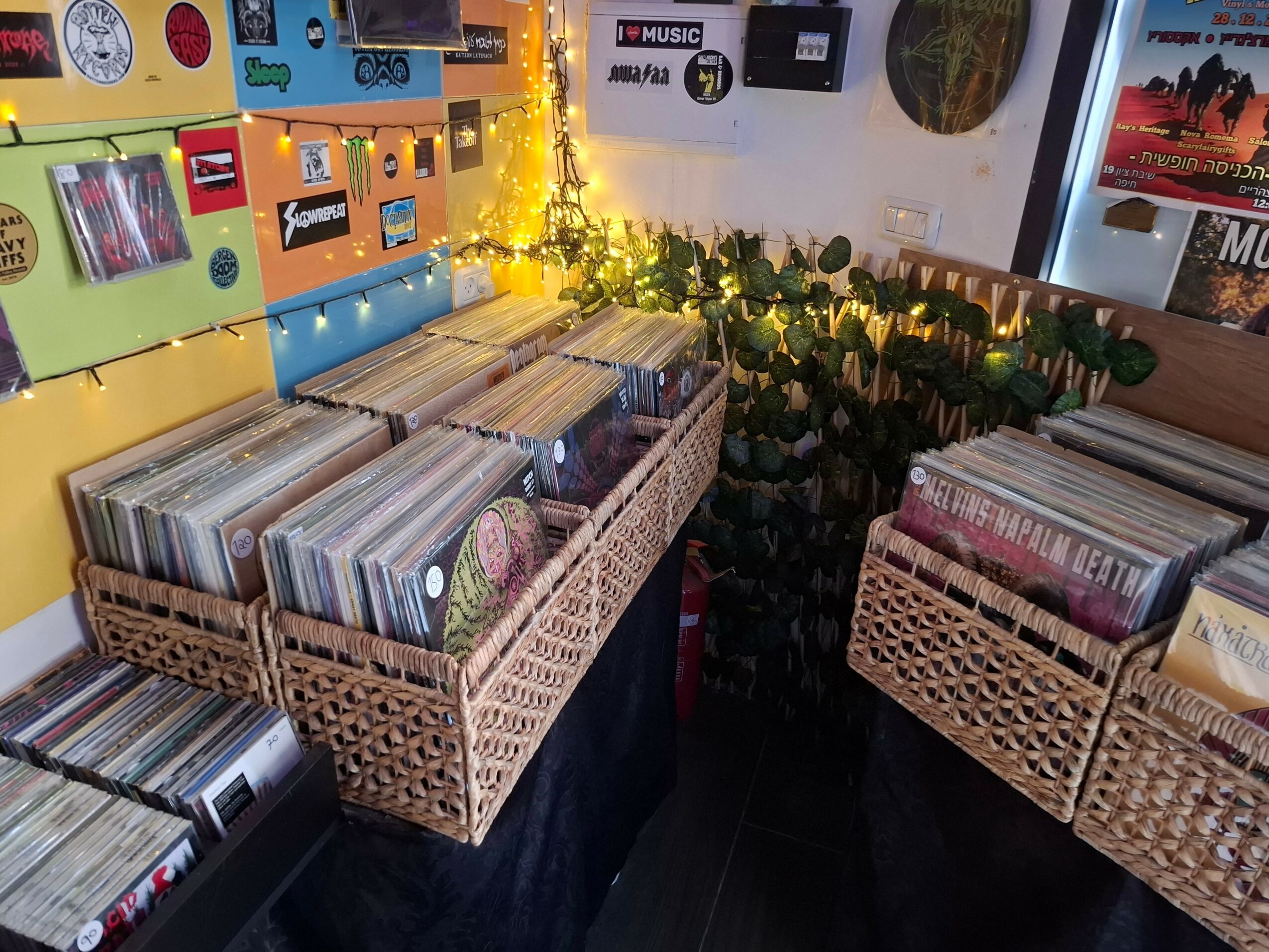 Three baskets filled with vinyl records on a table, with a wall decorated with posters and a string of lights behind them.