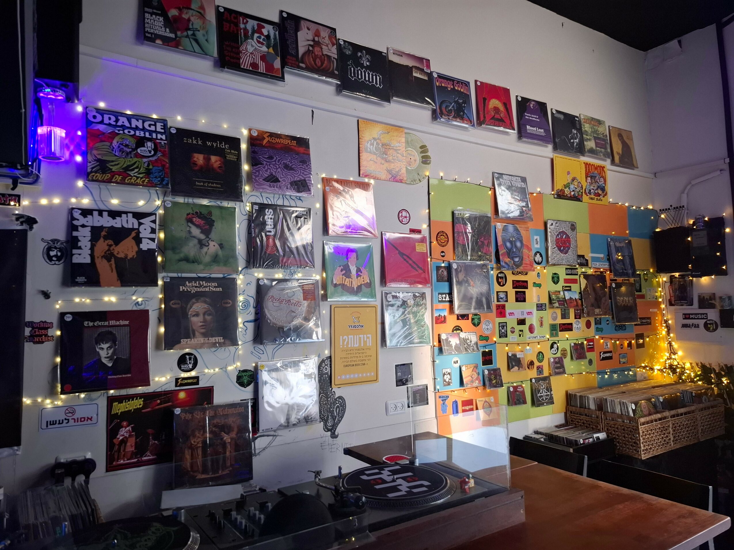Wall decorated with vinyl records, colorful album covers, and string lights, with a turntable and wooden table in a cozy room.