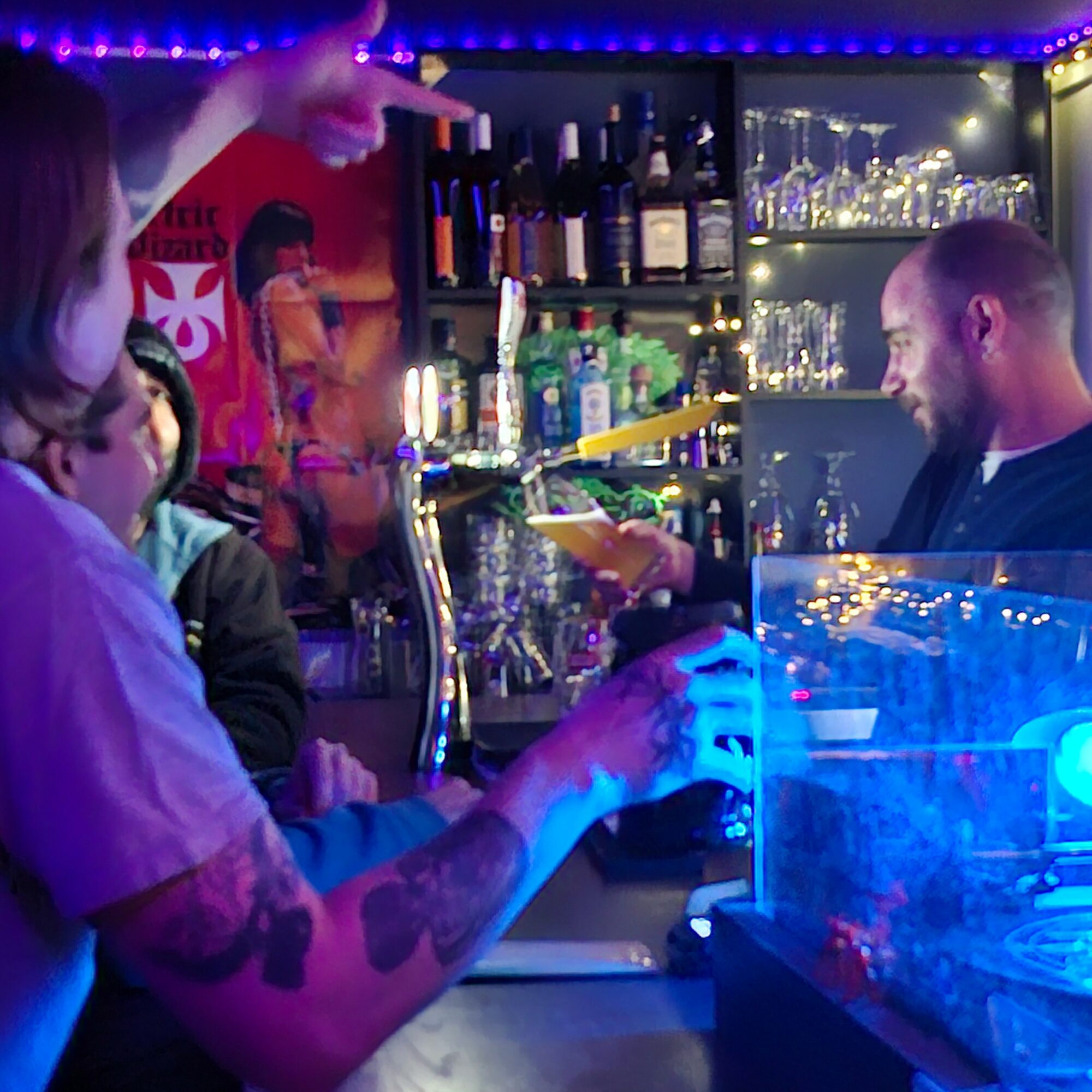 People at a bar with a bartender serving drinks, shelves with bottles and glasses in the background.
