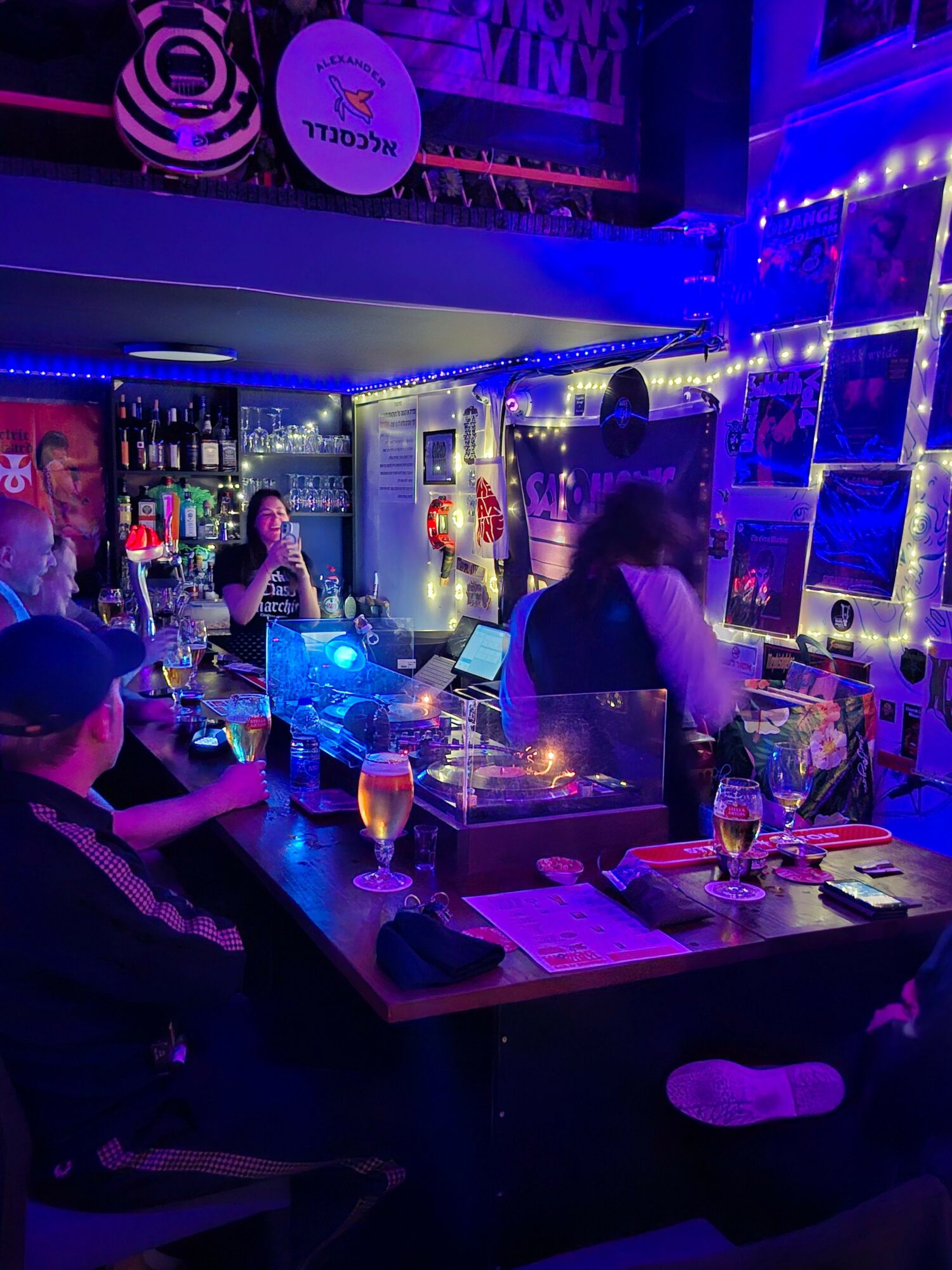 People gather around a bar with colorful bottles, illuminated by purple and blue lights, in a lively setting.