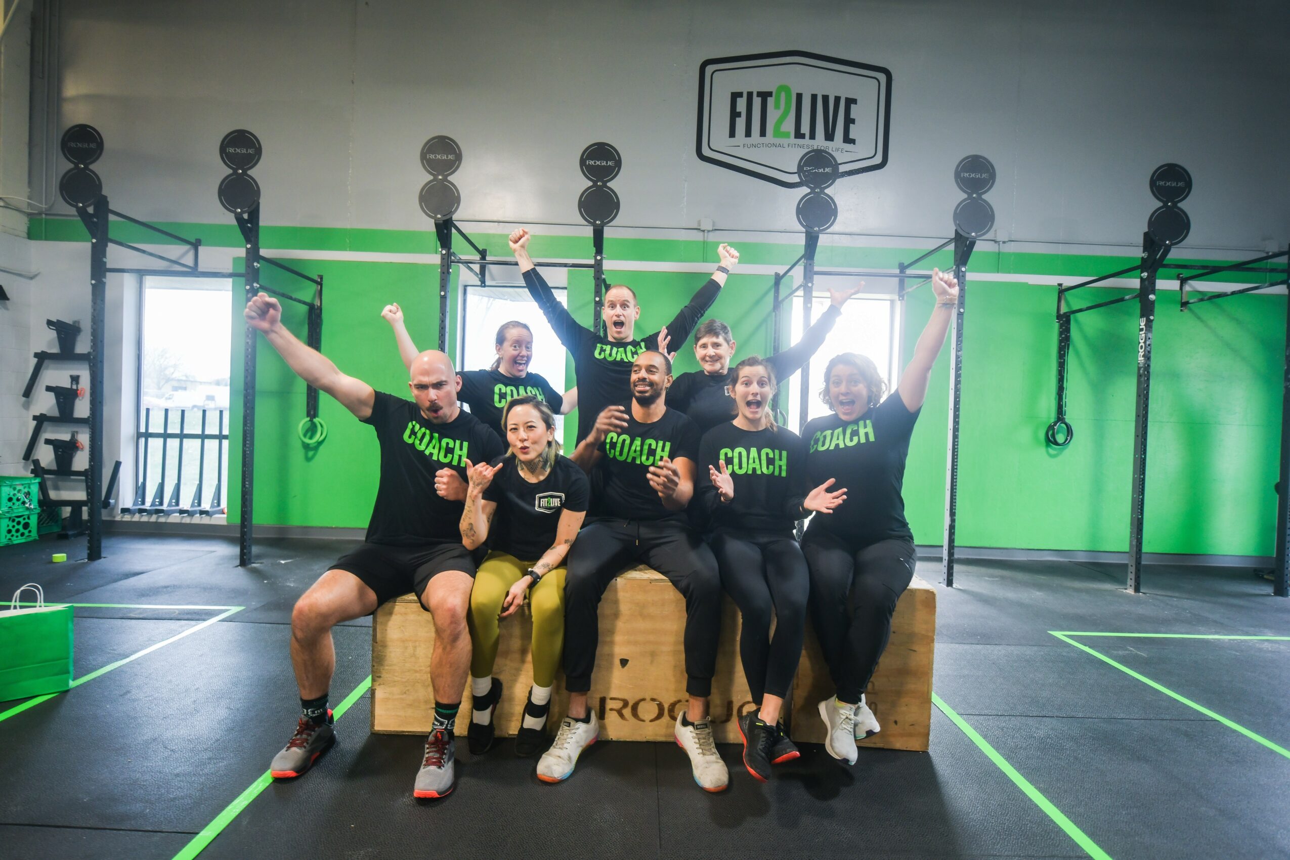 Group of six people sitting and standing in a gym, raising their arms, with a green wall and fitness equipment behind them.