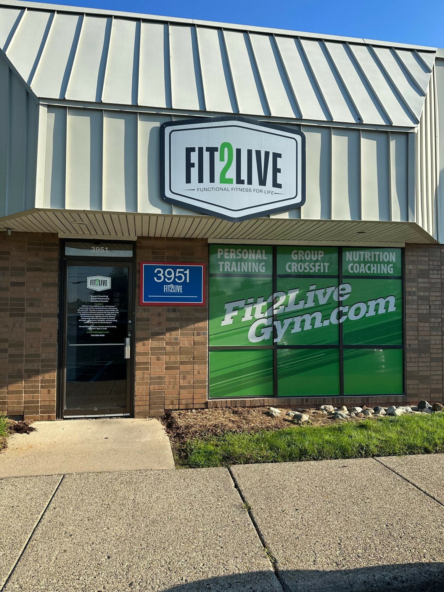 Fitness center sign and entrance with green promotional poster, brick wall, and sidewalk.