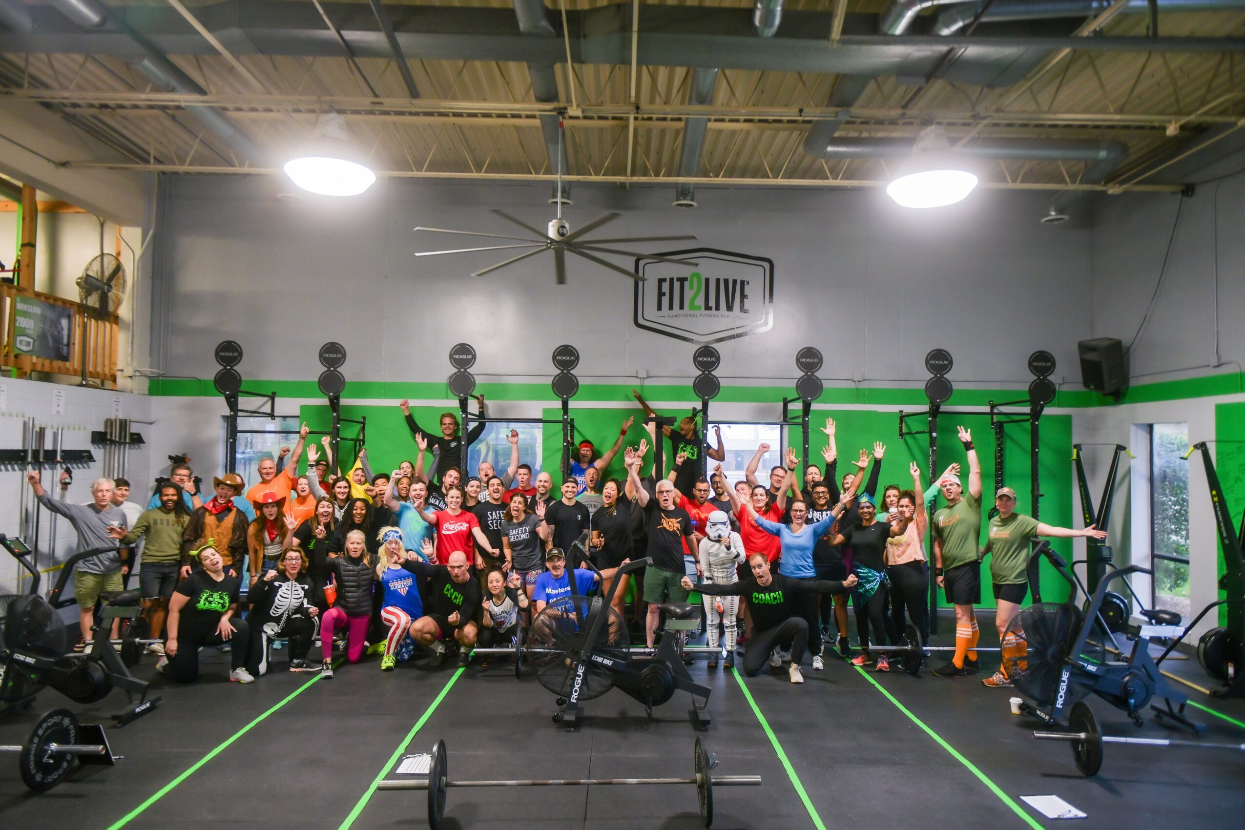 Group of people in a gym, some holding kettlebells overhead, with fitness equipment and a green wall in the background.