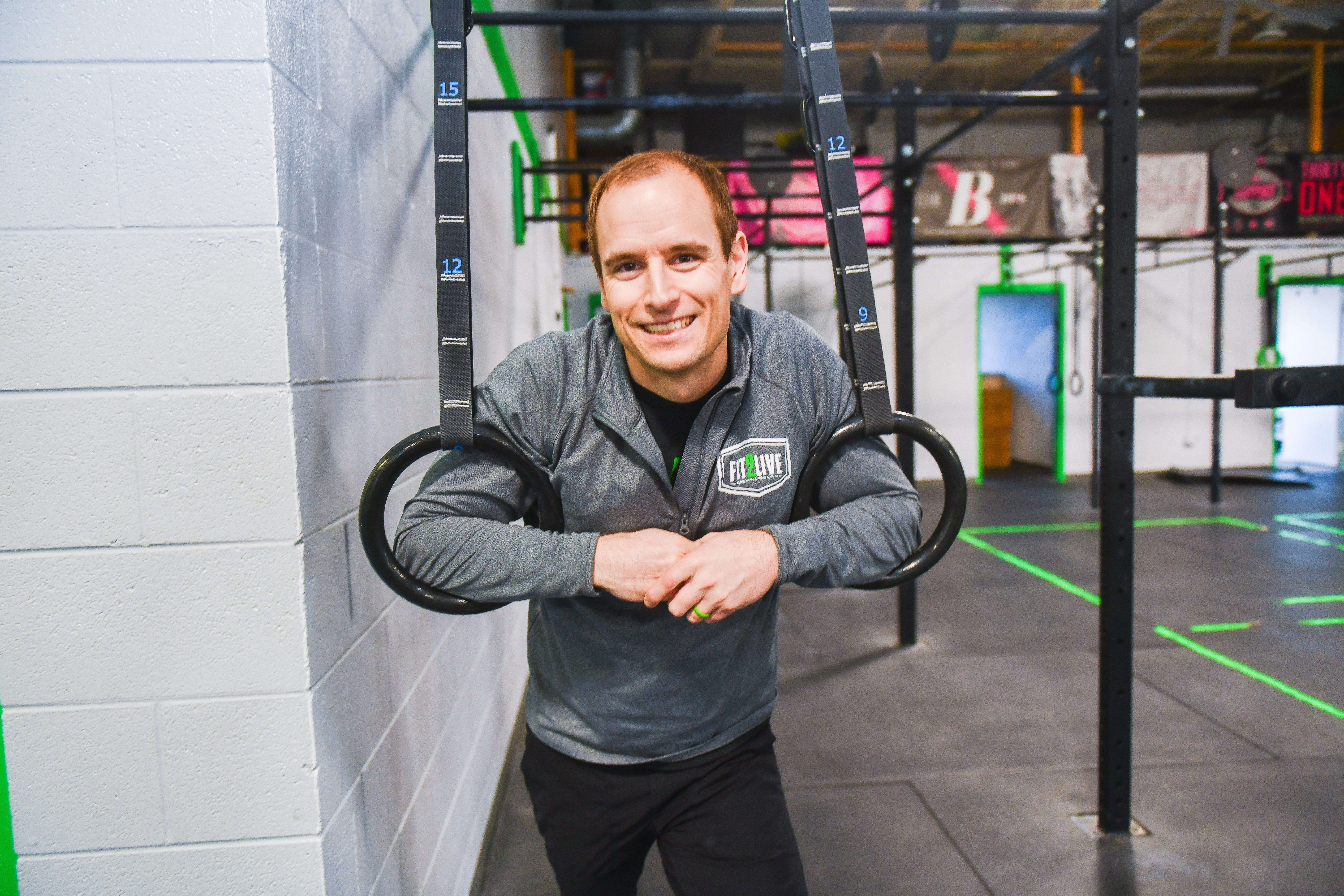 Smiling man leaning against a wall in a gym with workout equipment in the background.