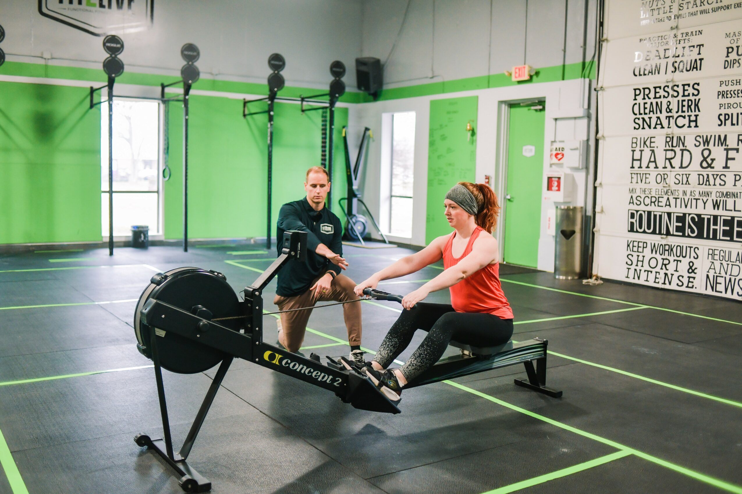 Two women using rowing machines in a gym with green and white walls, black equipment, and motivational posters, one assisting the other.