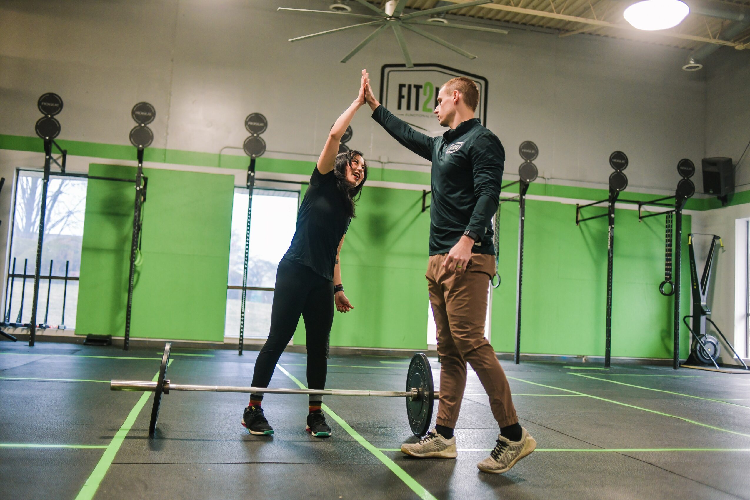 A woman and a man high-five in a gym with green walls and exercise equipment, both smiling and facing each other.