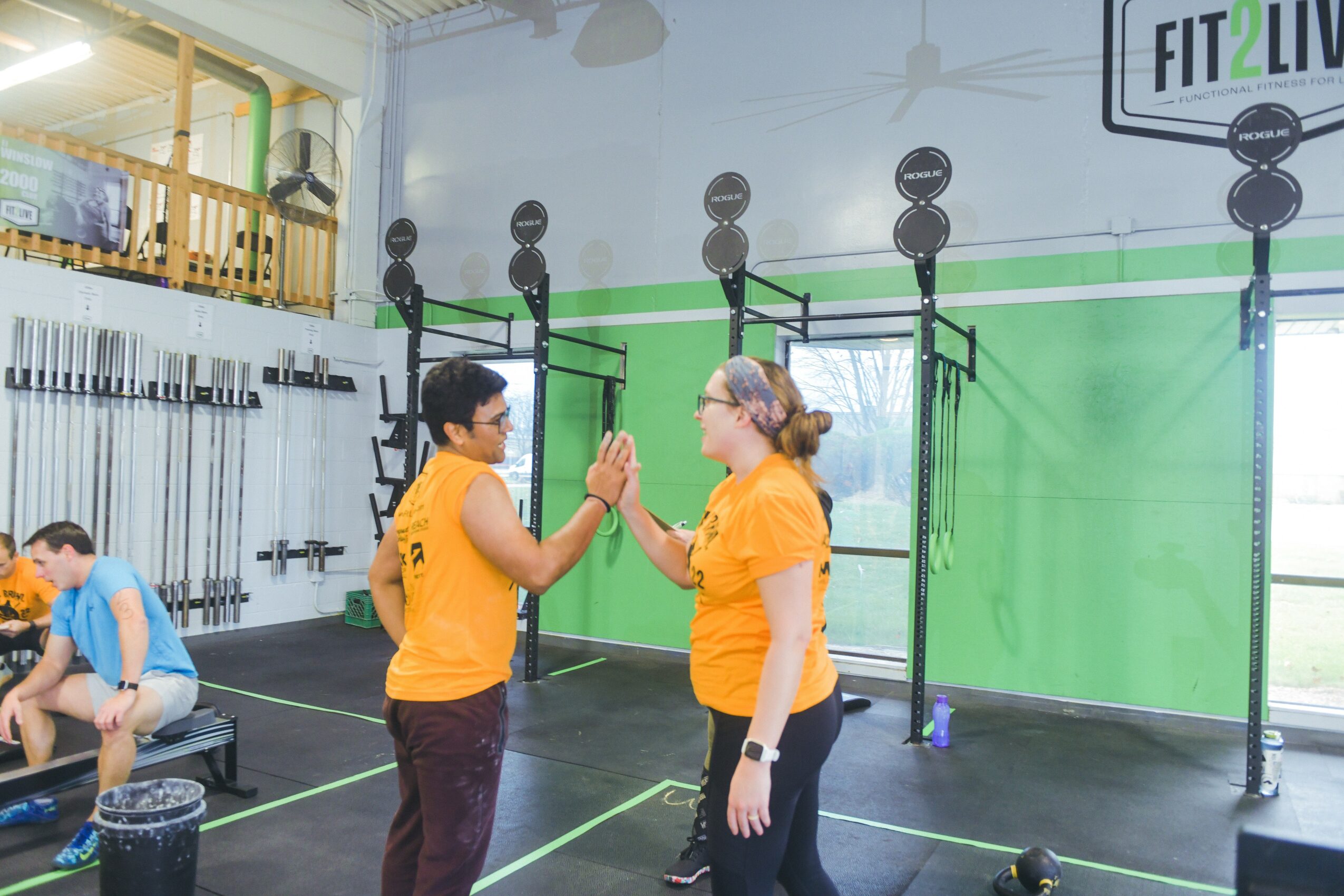 Two women in yellow shirts high-five in a gym with workout equipment and green walls.