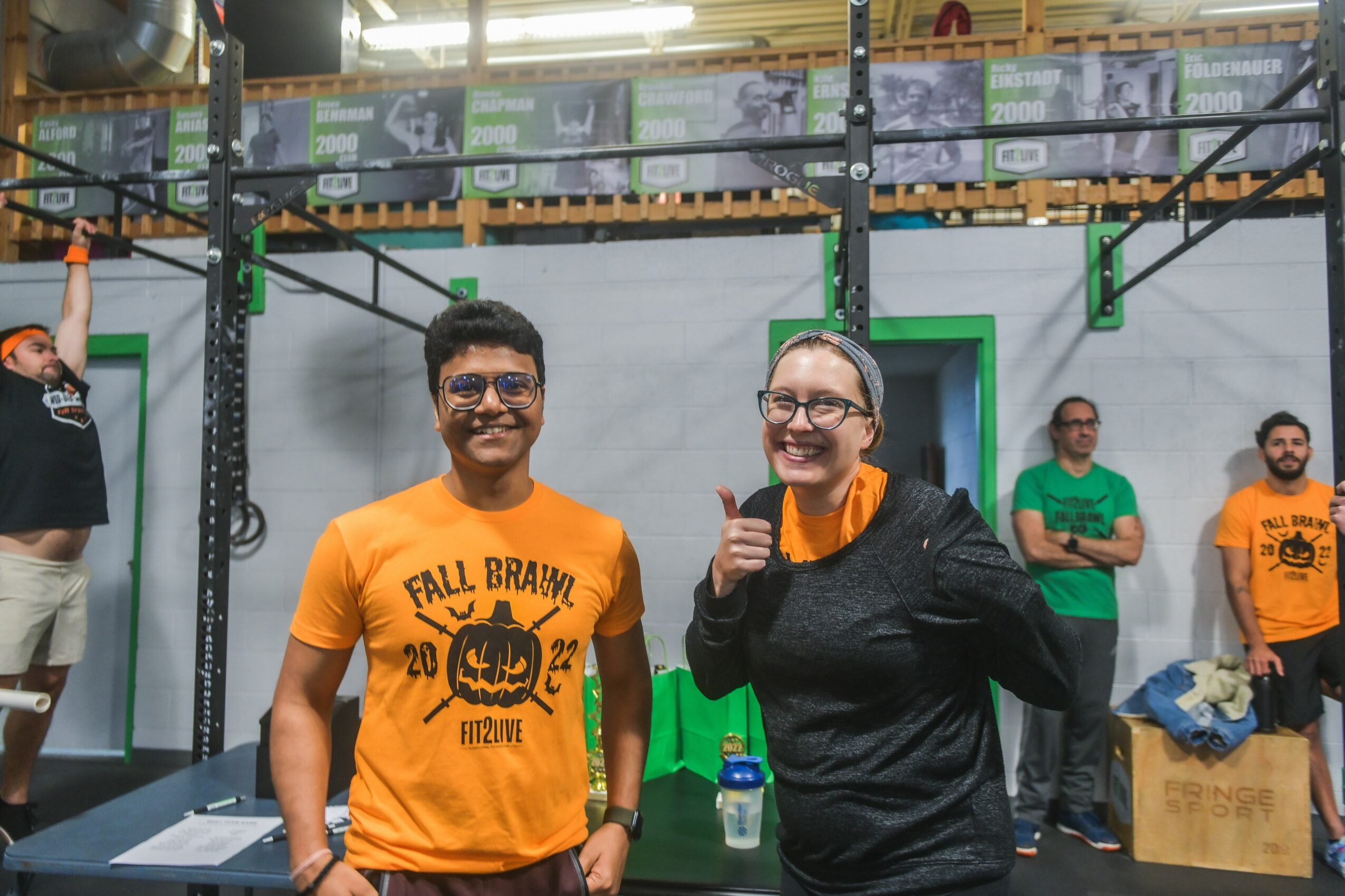 Group of people in a gym, two smiling individuals in the foreground, one giving a thumbs-up, others in the background, some wearing orange shirts.