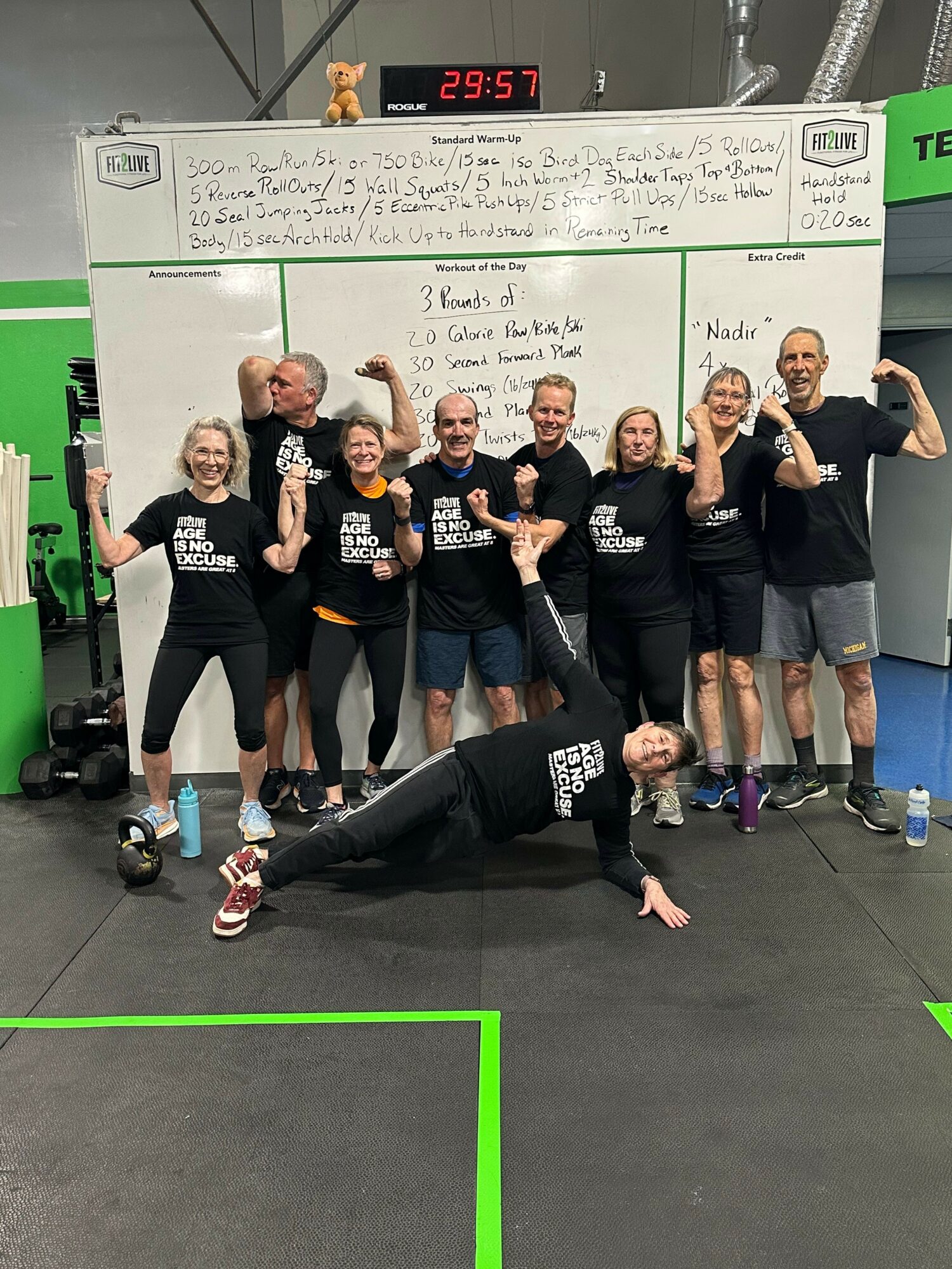 Group of eight people in a gym, some kneeling and some standing, giving thumbs-up gestures, with a whiteboard in the background.