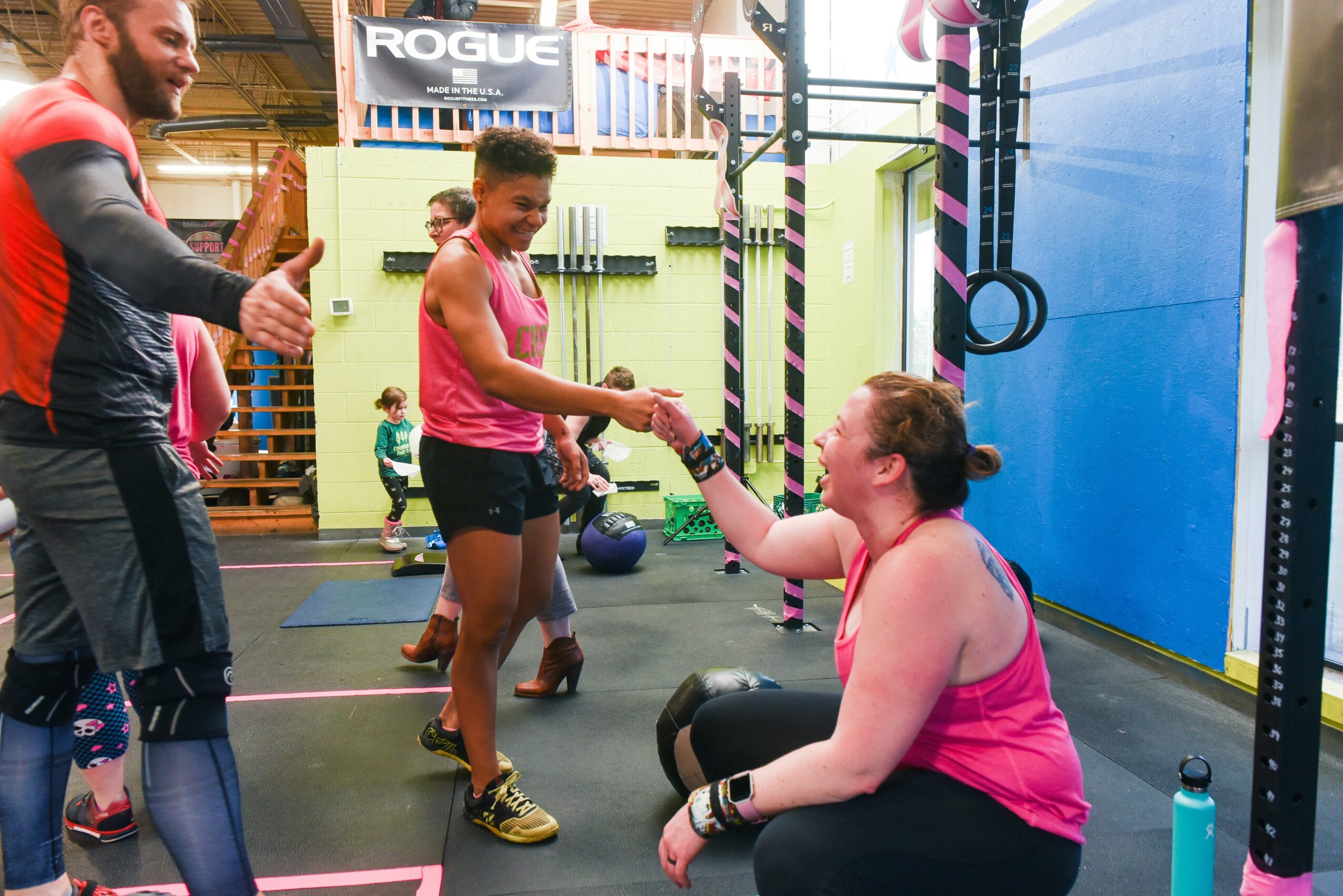 Two women shake hands in a gym with fitness equipment and children in the background, one woman sitting and the other standing.