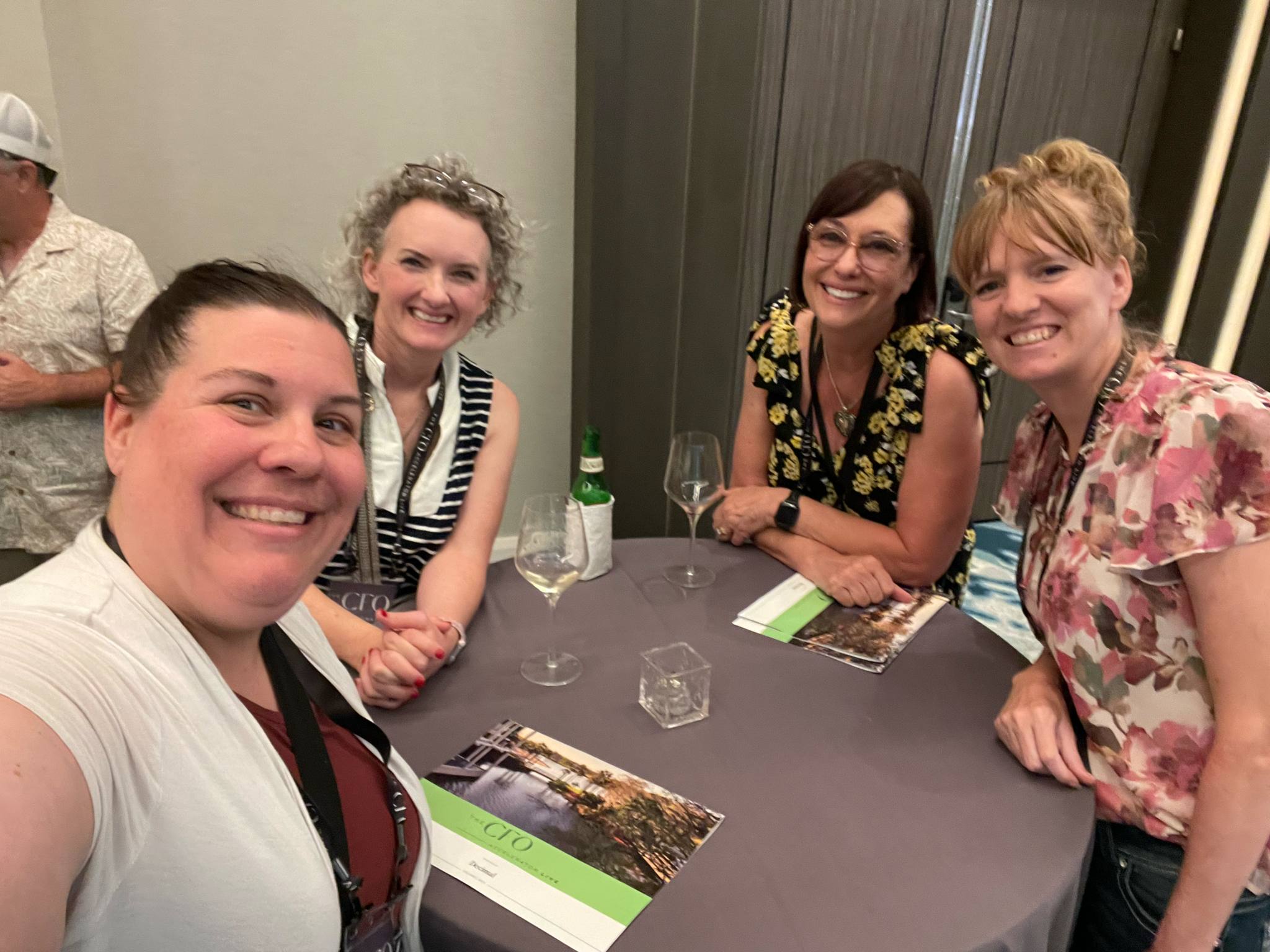 Four women sitting around a table smiling at the camera, with drinks and brochures on the table.