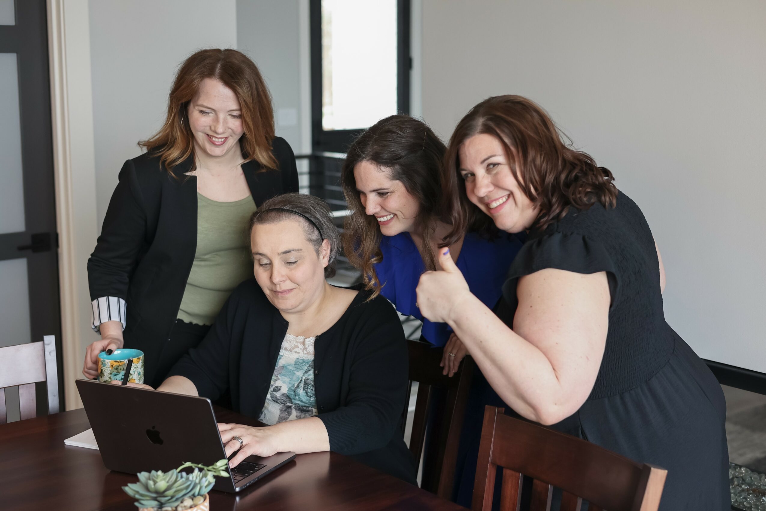 Four women gathered around a laptop, smiling and looking at the screen in a room with a window.