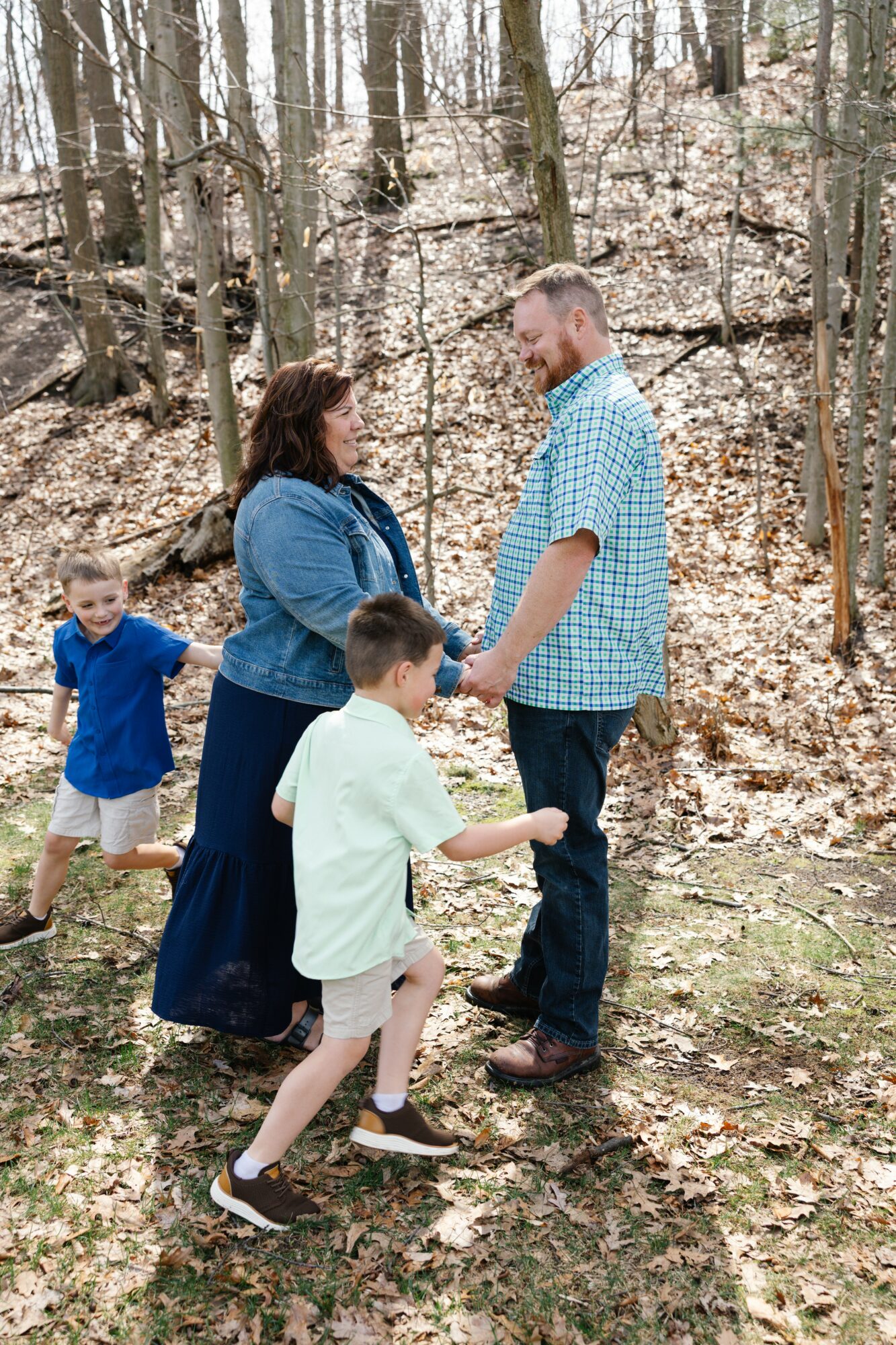 A family of four holding hands in a wooded area with leaf-covered ground, trees, and a slope in the background.