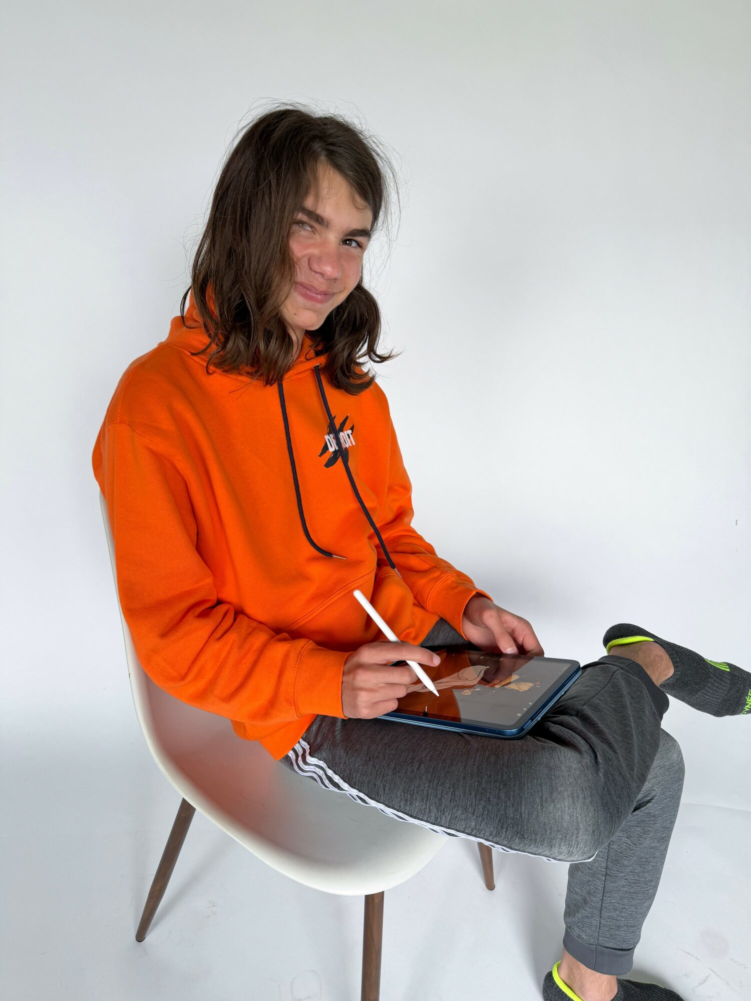 Person with shoulder-length brown hair wearing an orange hoodie, sitting on a white chair, using a tablet.