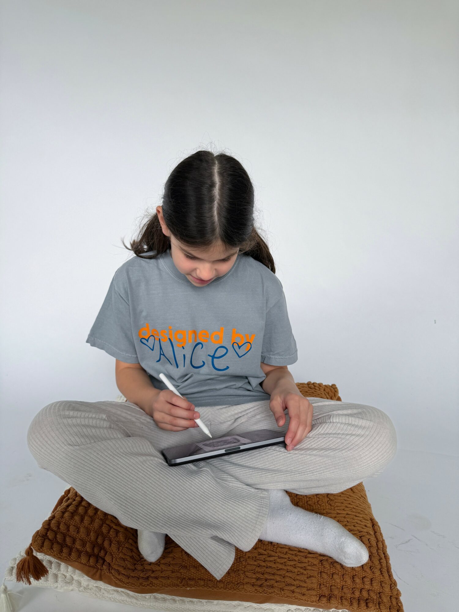 Child sitting cross-legged on a brown blanket, looking down at a tablet, against a plain light background.