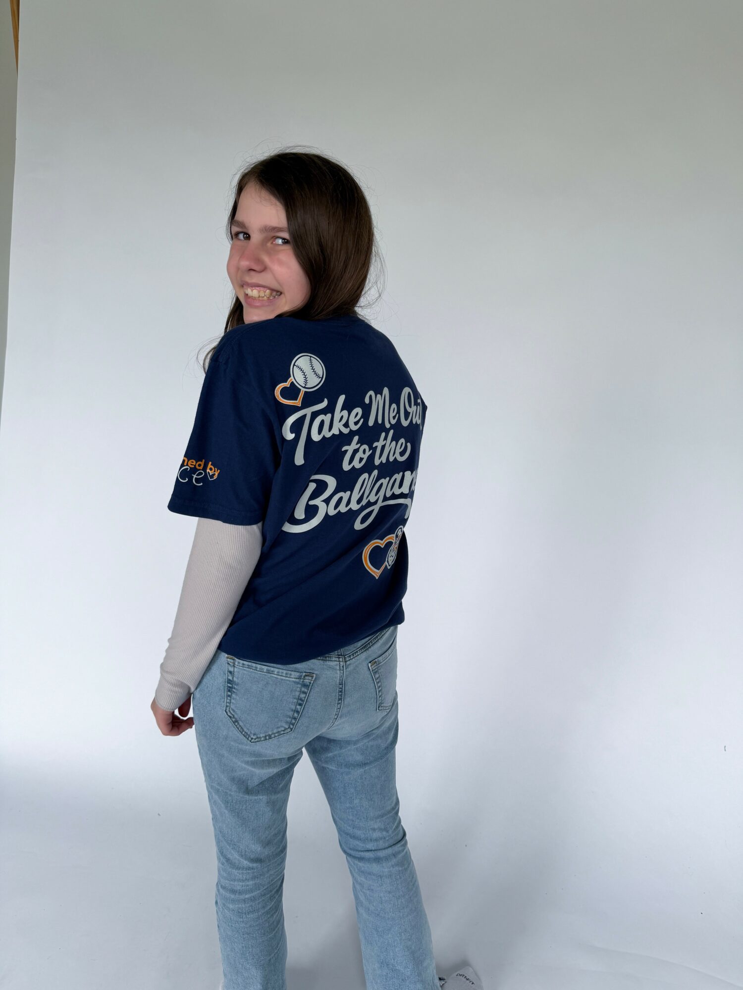 Young woman with long brown hair smiling, wearing a navy blue T-shirt and light blue jeans, standing against a plain white background.
