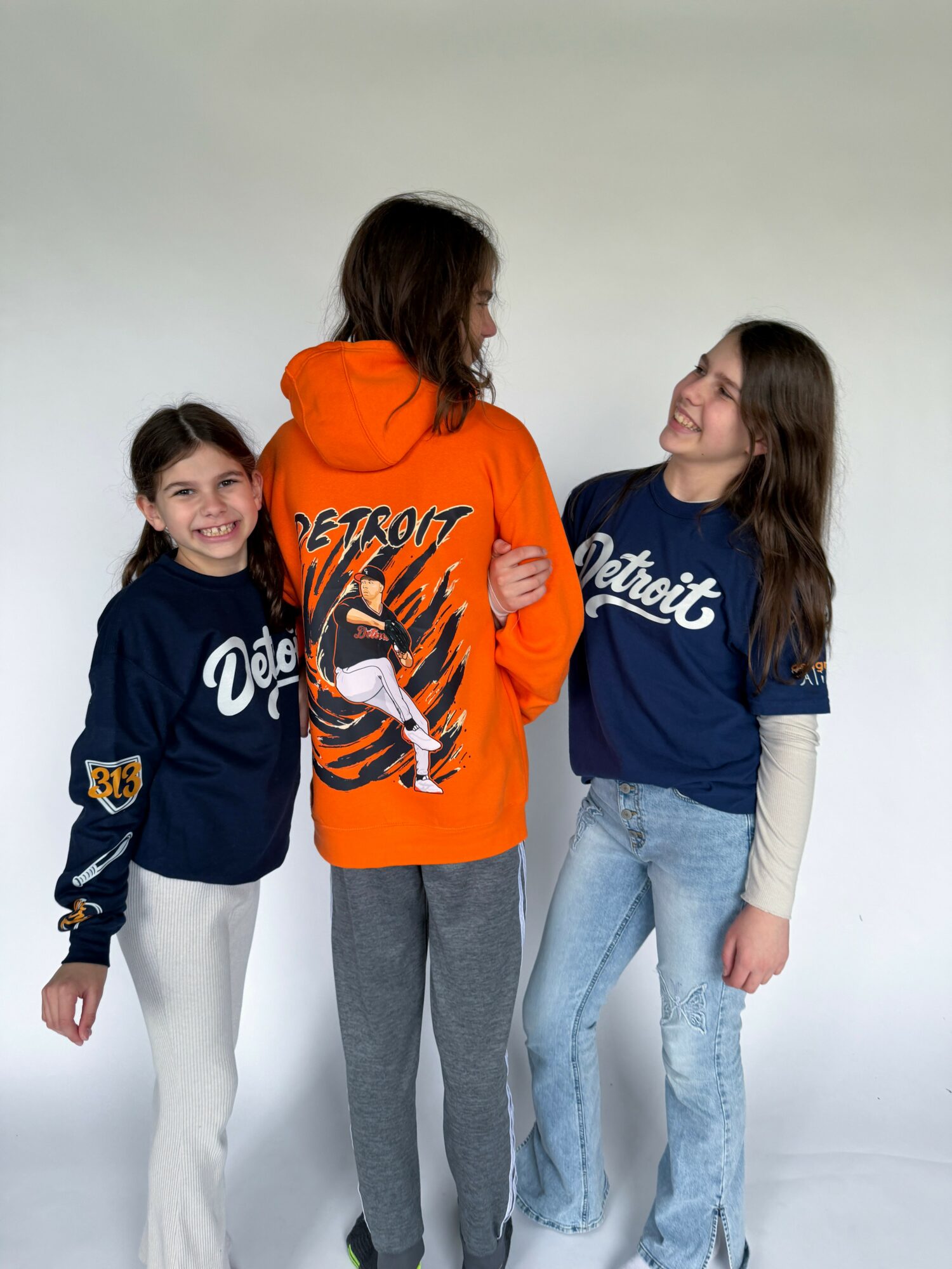 Three children standing together, two girls wearing Detroit sports jerseys and a boy in an orange hoodie with a tiger graphic, smiling.