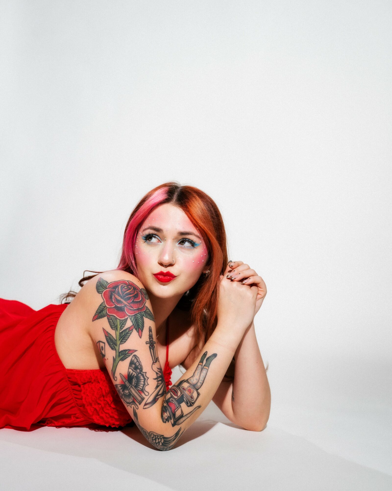Young woman with red hair and tattoos lying on her side, wearing a red dress, against a white background.