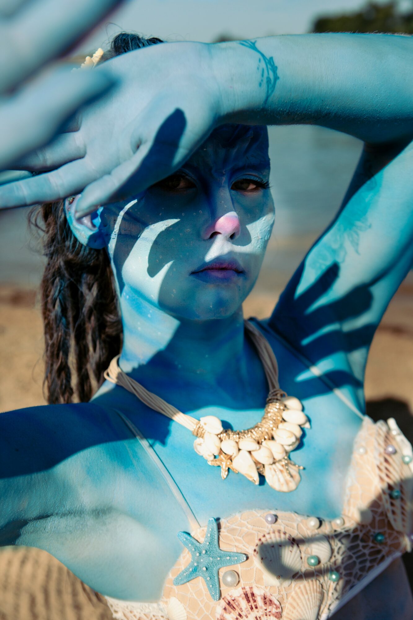 Woman with blue body paint, wearing a shell necklace and starfish, shielding her eyes with her arm, outdoors.