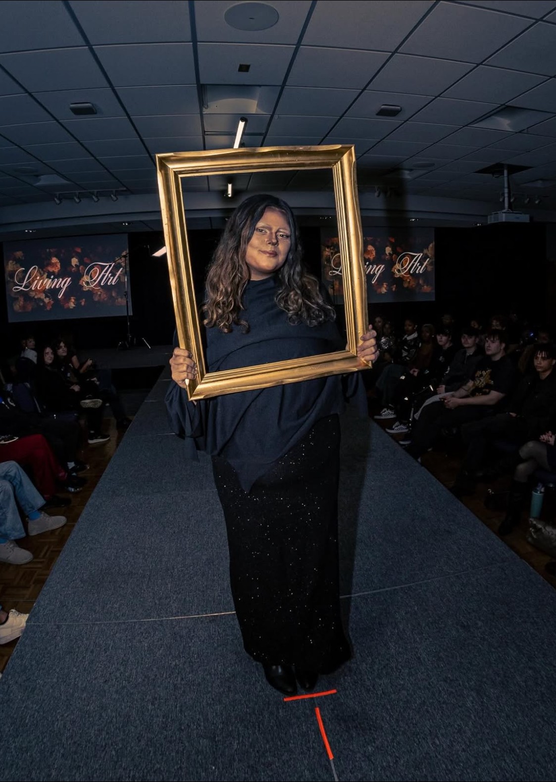 Person walking on runway holding a large empty gold frame, audience seated on both sides, dark ceiling with lights.