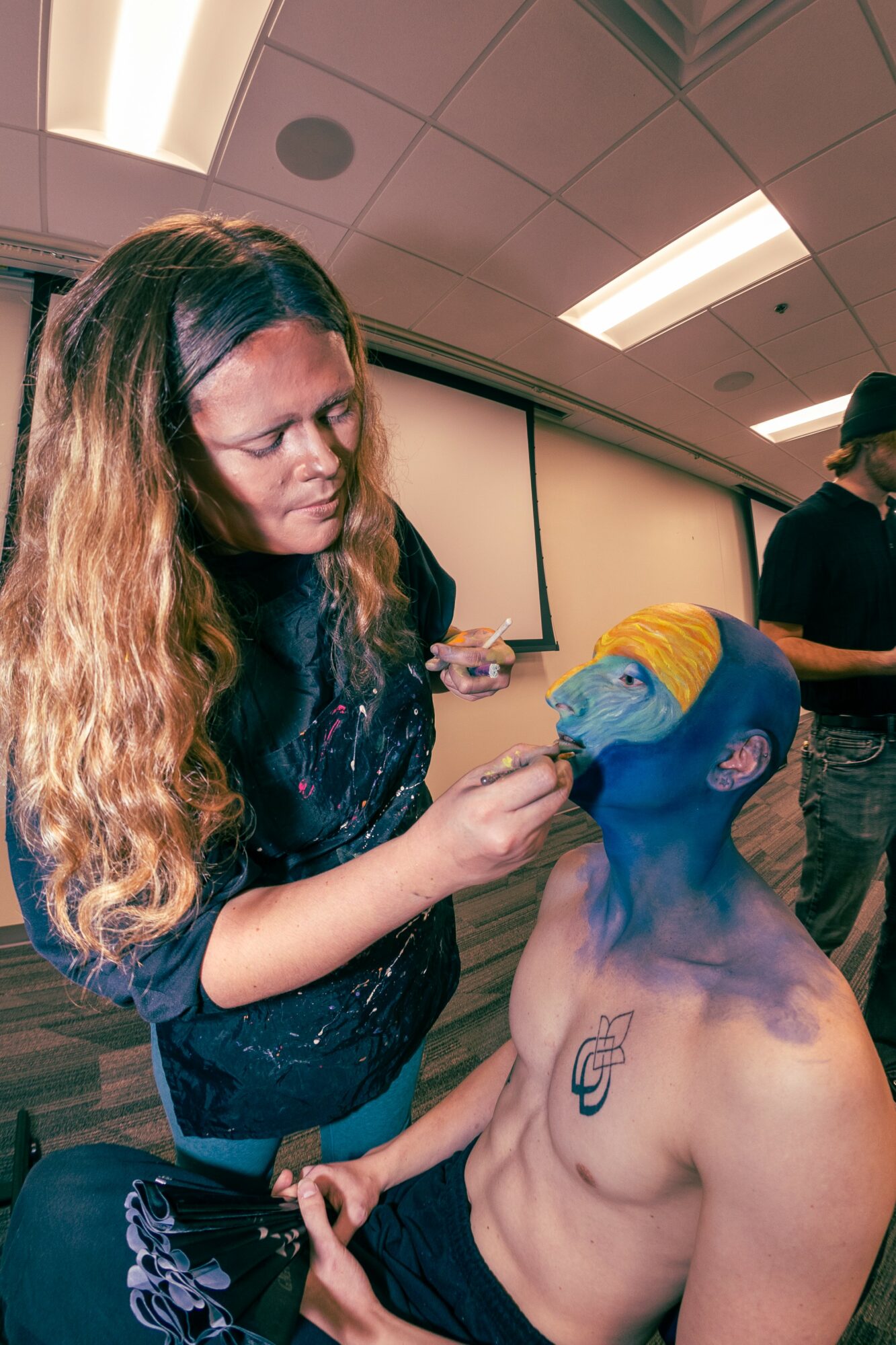 Woman painting face of a shirtless person with blue and yellow face paint, in a room with ceiling lights.