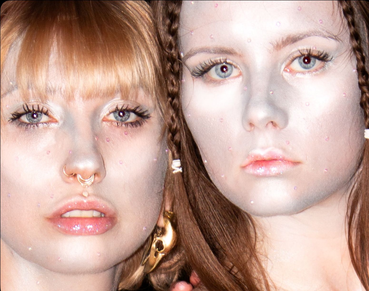 Two women with light skin and light-colored eyes, lying down close together, with one woman having a nose ring and gold earring.