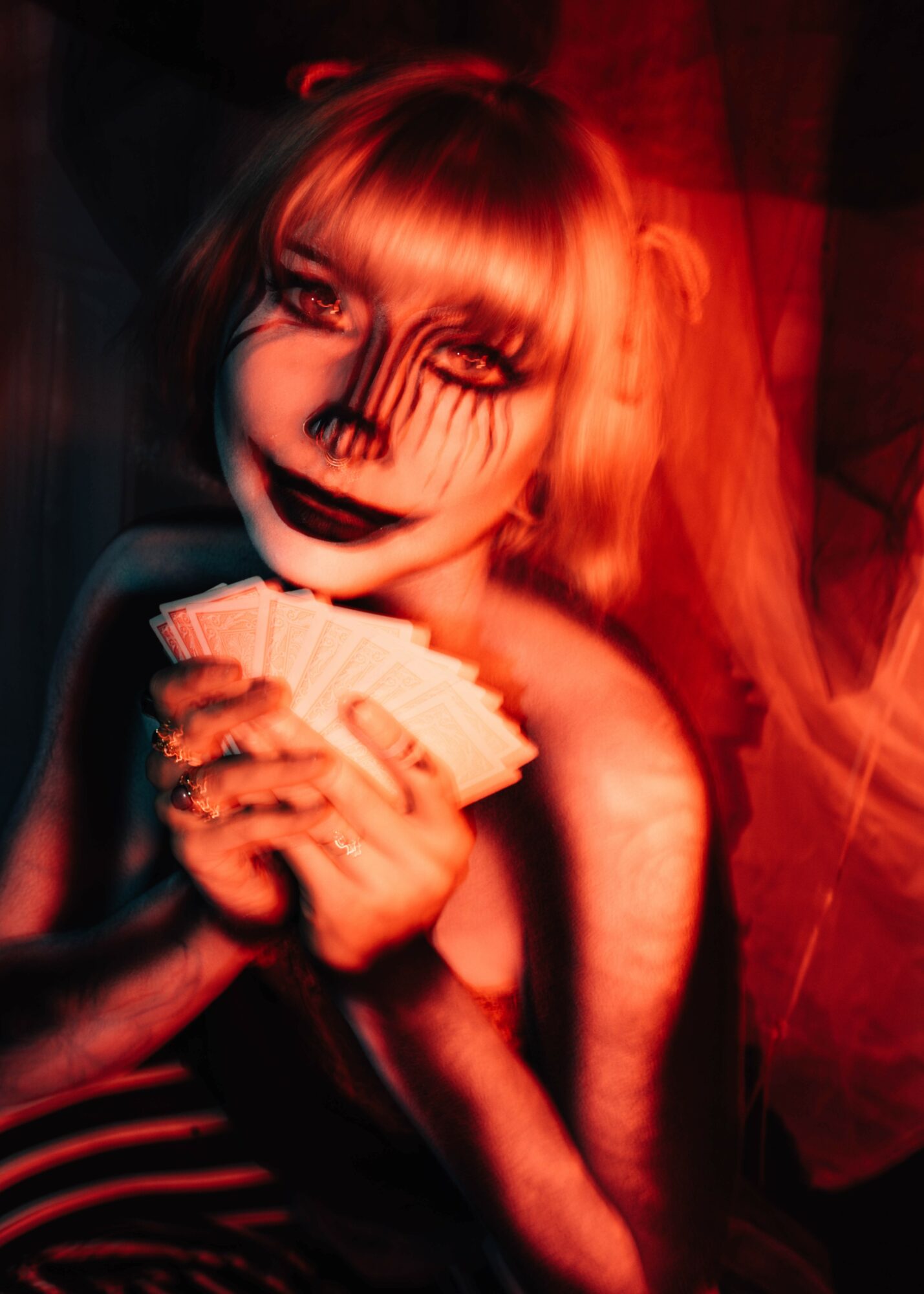 Person with face painted as a skeleton, holding a stack of cards, in a dimly lit setting with red lighting.