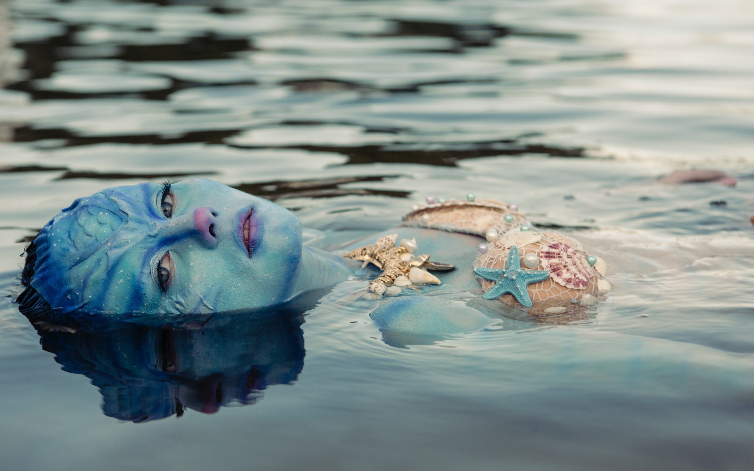 Person with blue face paint and a swim cap floats in water near starfish and seashells, looking at the camera.