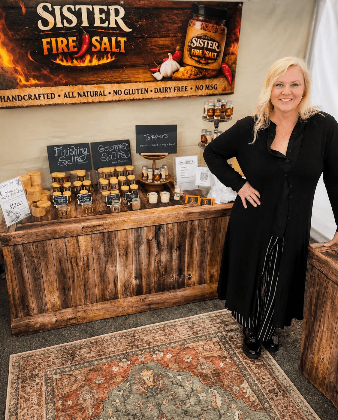 Woman with blonde hair in black coat standing next to a wooden display table with jars of Sister Fire Salt, a chalkboard sign, and product labels, in a store setting.