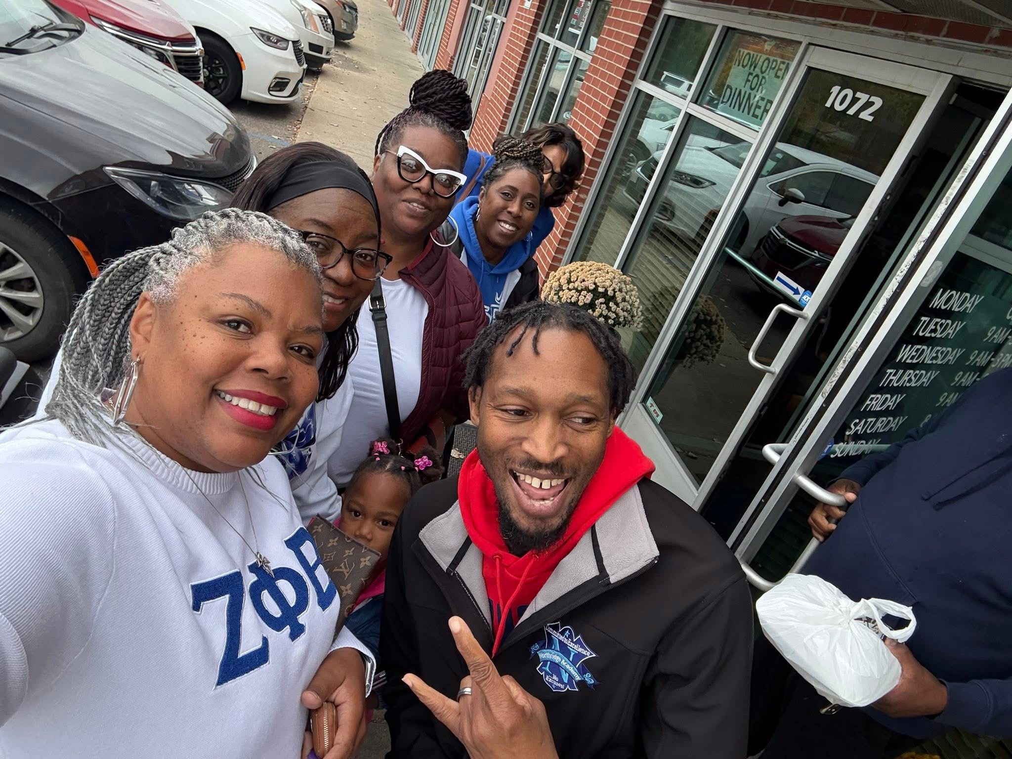 Group of six people smiling outside a building, some making peace signs, with cars parked nearby.