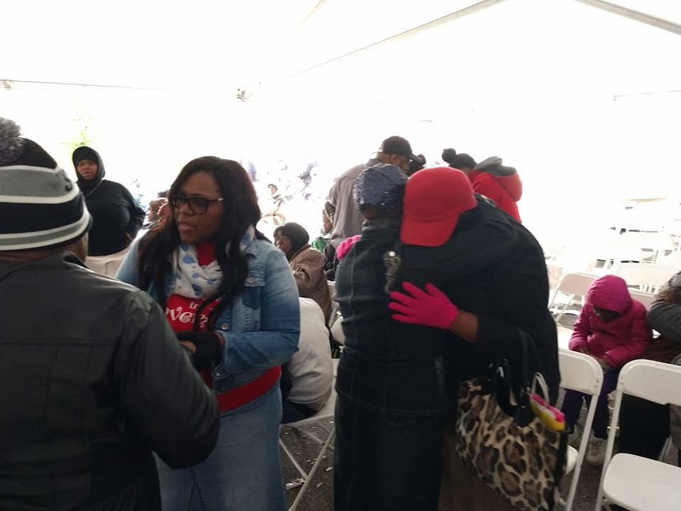 People gathered outdoors under a white canopy, some hugging, others sitting on chairs, dressed warmly with hats and gloves.