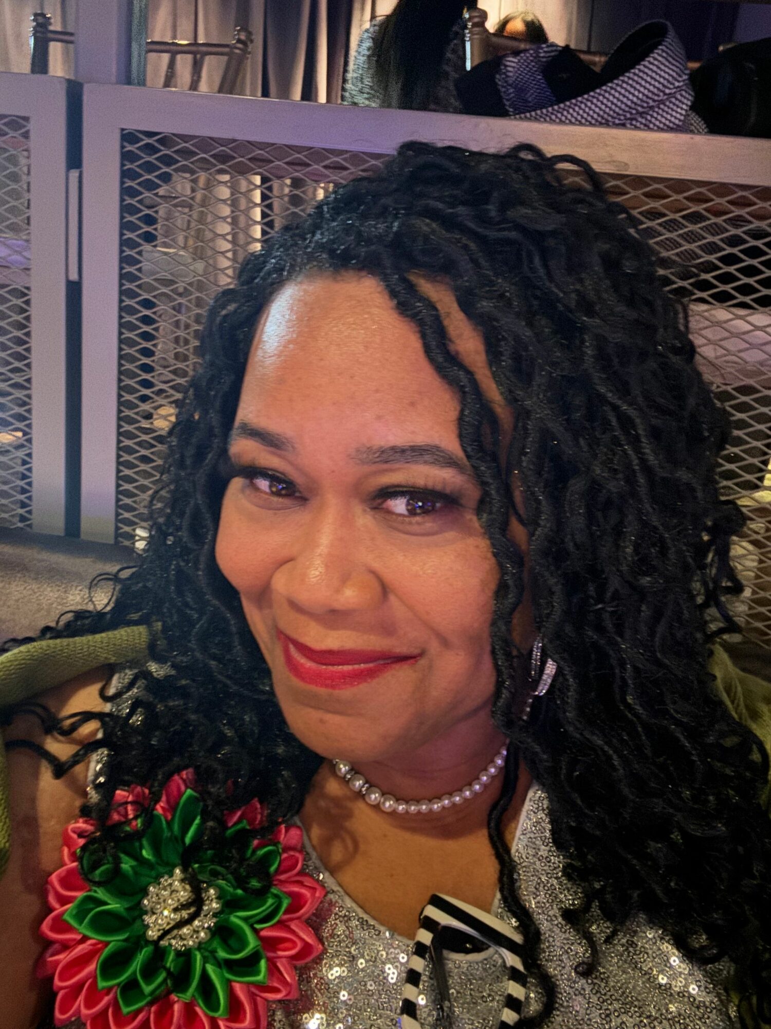 Woman with curly hair, smiling, wearing a pearl necklace and a decorative flower accessory, in a close-up portrait.