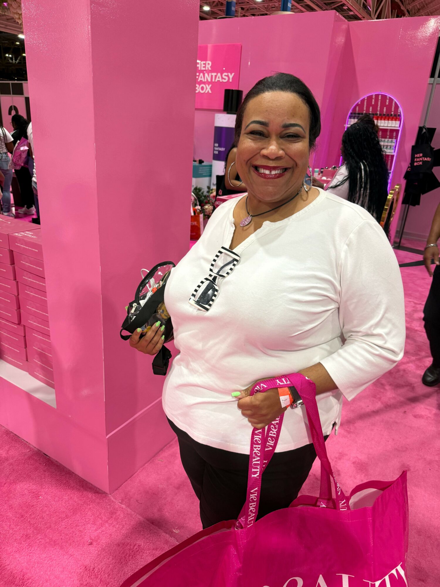 Woman smiling at a pink booth with beauty products, holding a pink shopping bag, in a store or event setting.