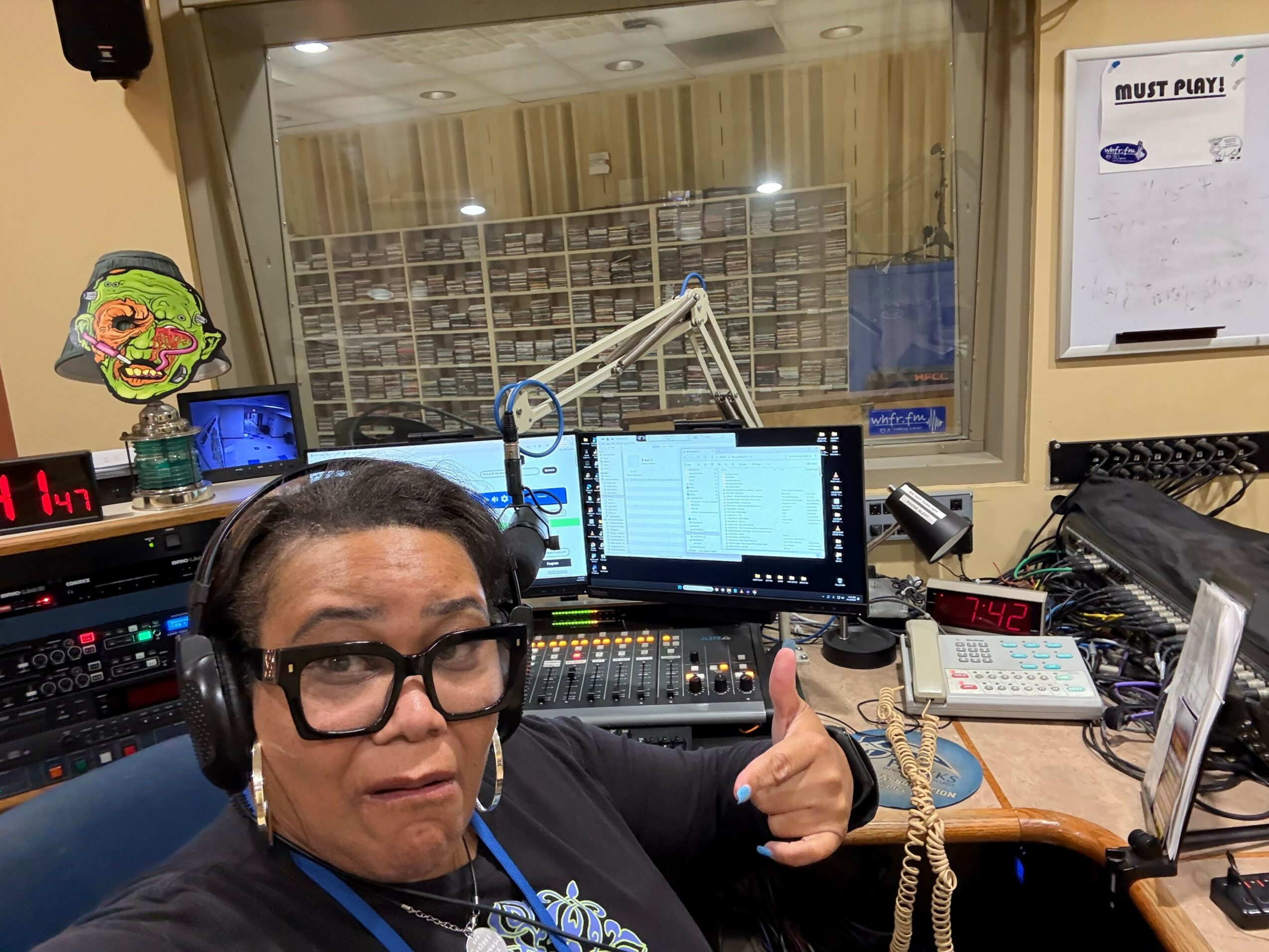Person wearing headphones and glasses in a radio studio with equipment and a computer monitor, smiling at the camera.