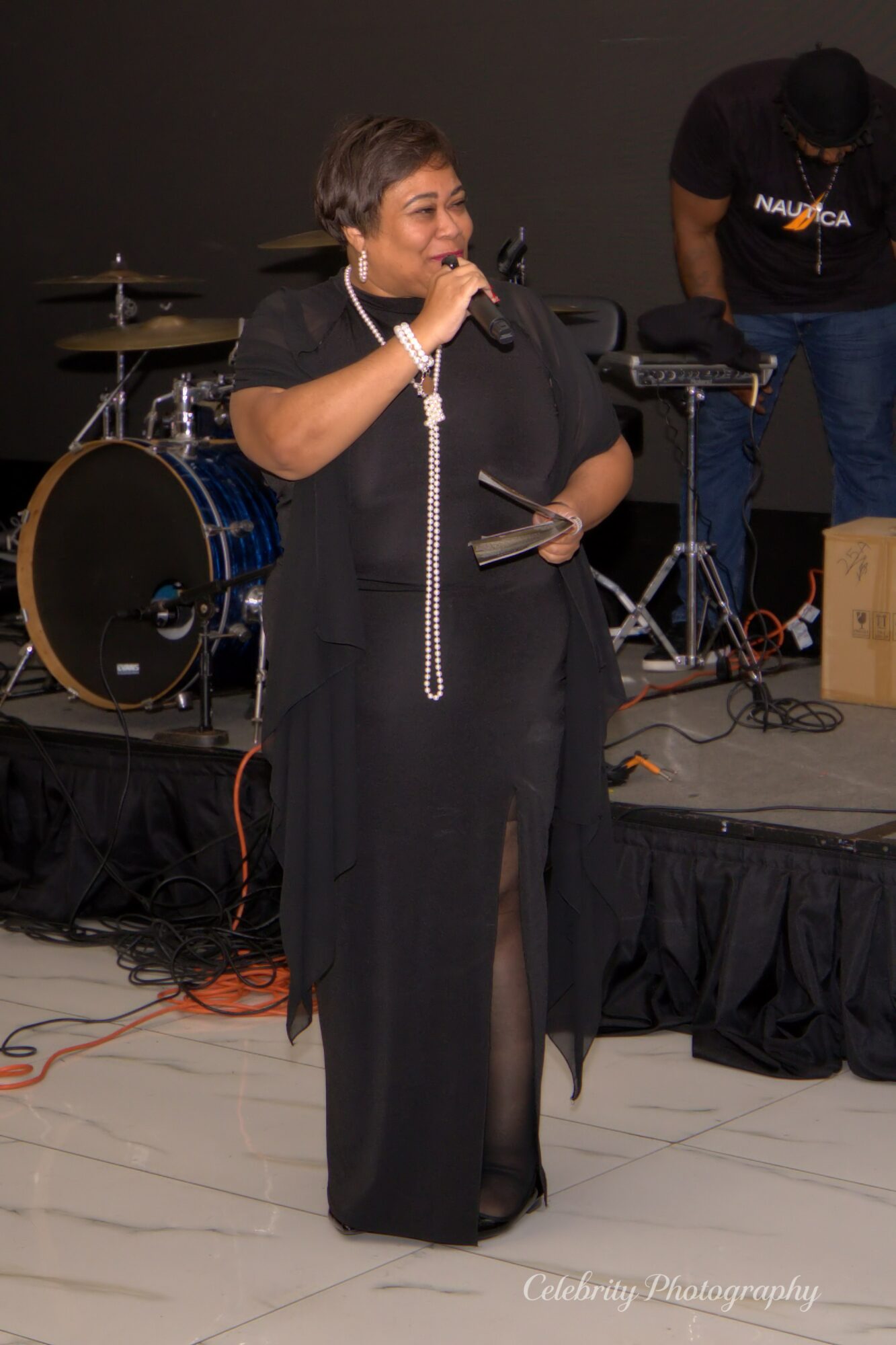 Woman speaking into microphone on stage with musical instruments in background, wearing black outfit, holding a tablet.