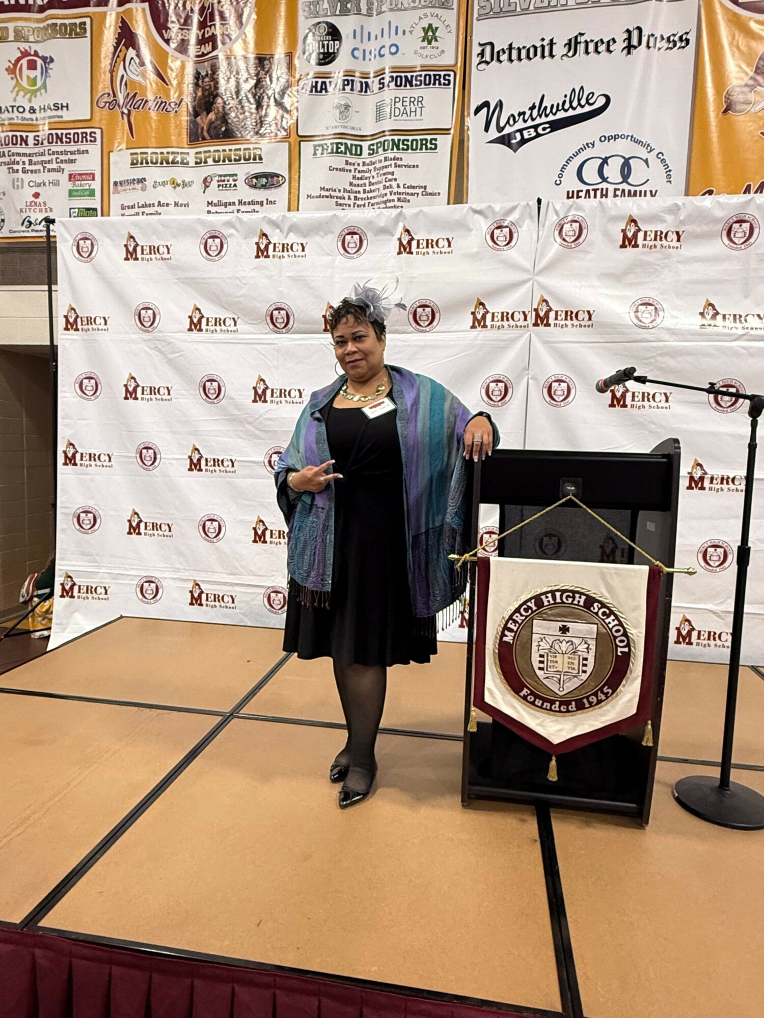 Woman standing on stage with a trophy, backdrop with logos, and a banner, holding a certificate, in a formal event.