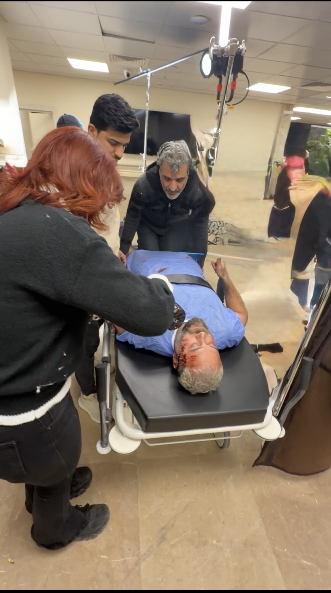 Medical team attending to a person lying on a hospital bed, with others in the background in a clinical setting.