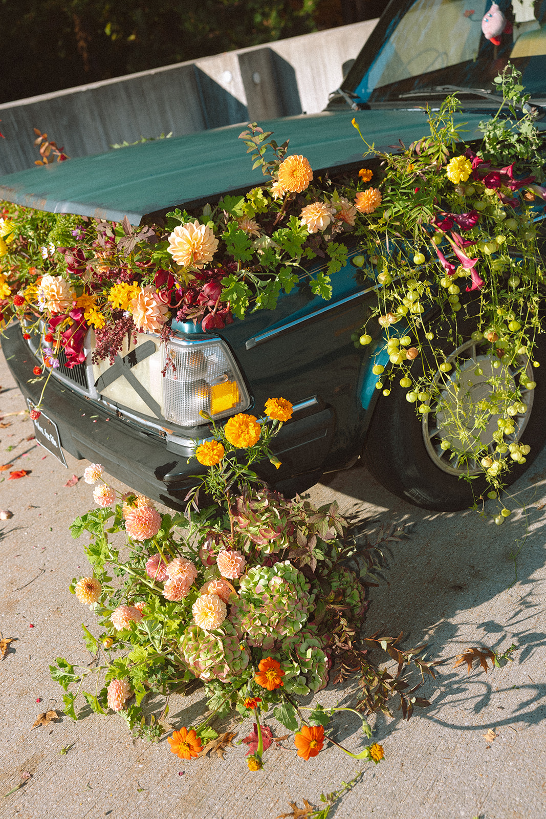 Car decorated with colorful flowers and greenery, with flowers arranged on the trunk and a bouquet on the ground nearby.