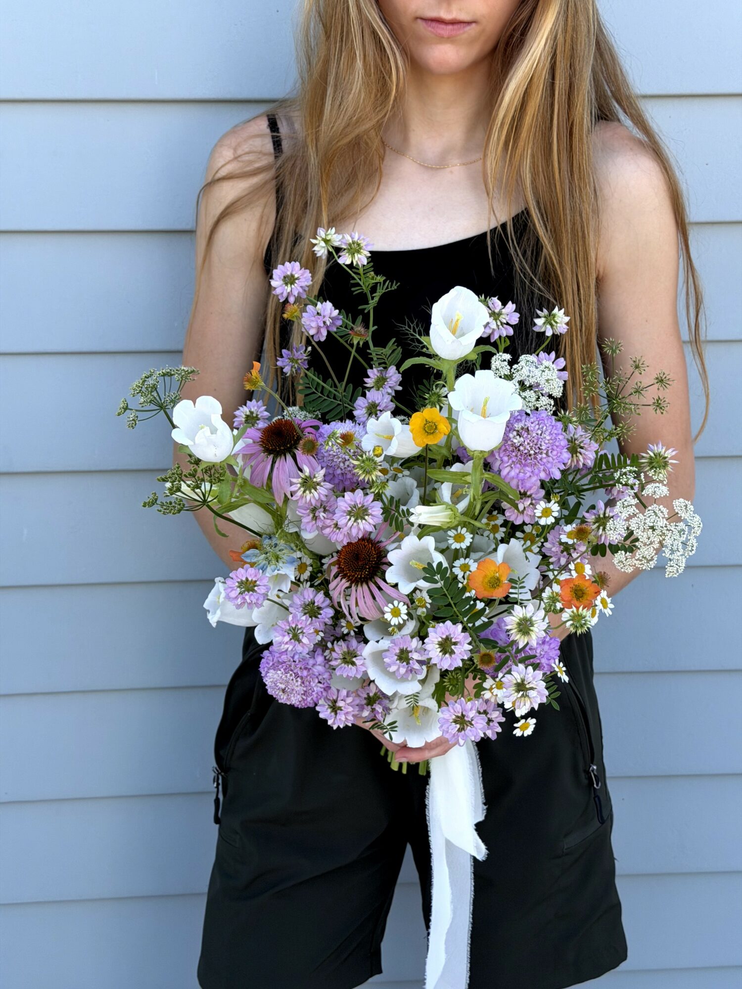 Person holding a large bouquet of mixed flowers, standing against a light blue wall, wearing a black top and shorts.