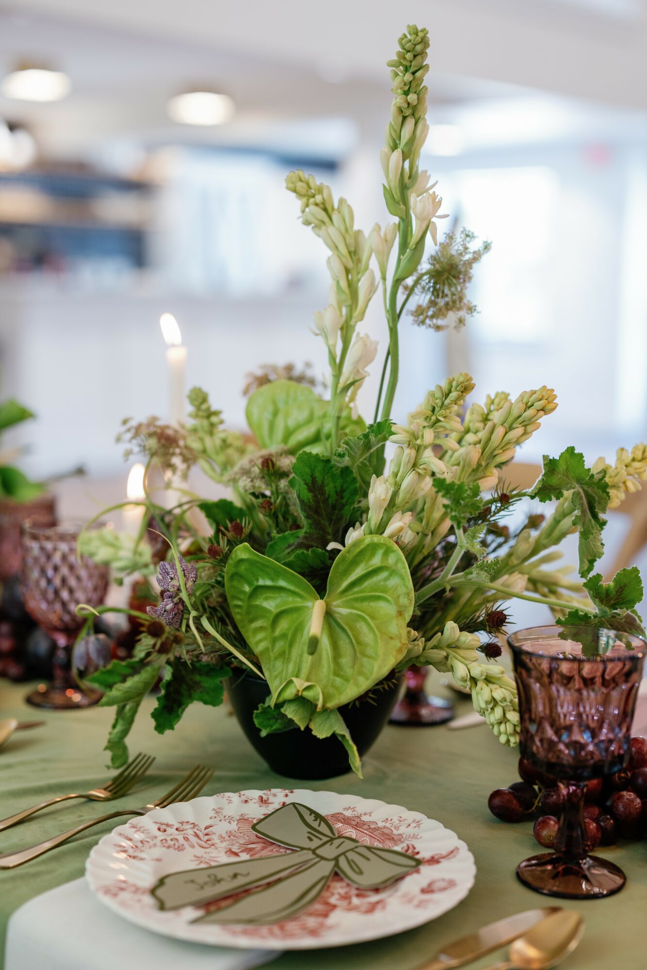 Flower arrangement with green leaves and tall stalks in a black vase on a table, with a decorative plate and purple glasses nearby.