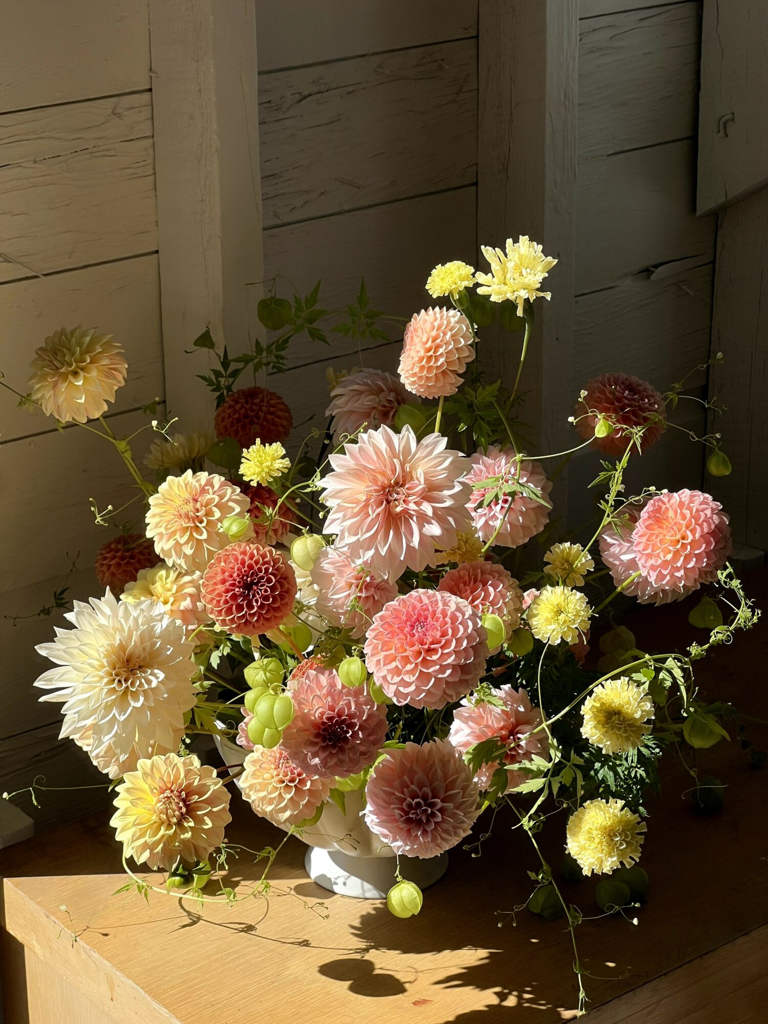 Flower arrangement with pink, yellow, and white blooms in a white vase on a wooden surface, against a wooden wall background.