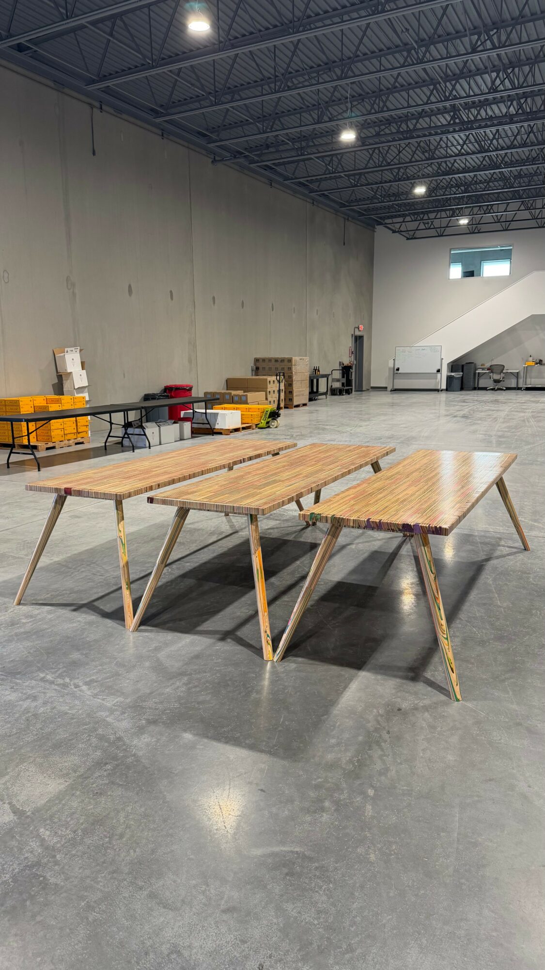 Two wooden tables with angled legs in a large industrial space with a high ceiling and concrete floor.