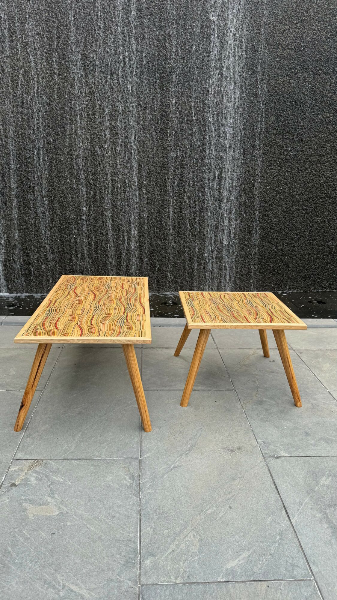 Two wooden tables with angled legs on a tiled floor against a dark textured wall.