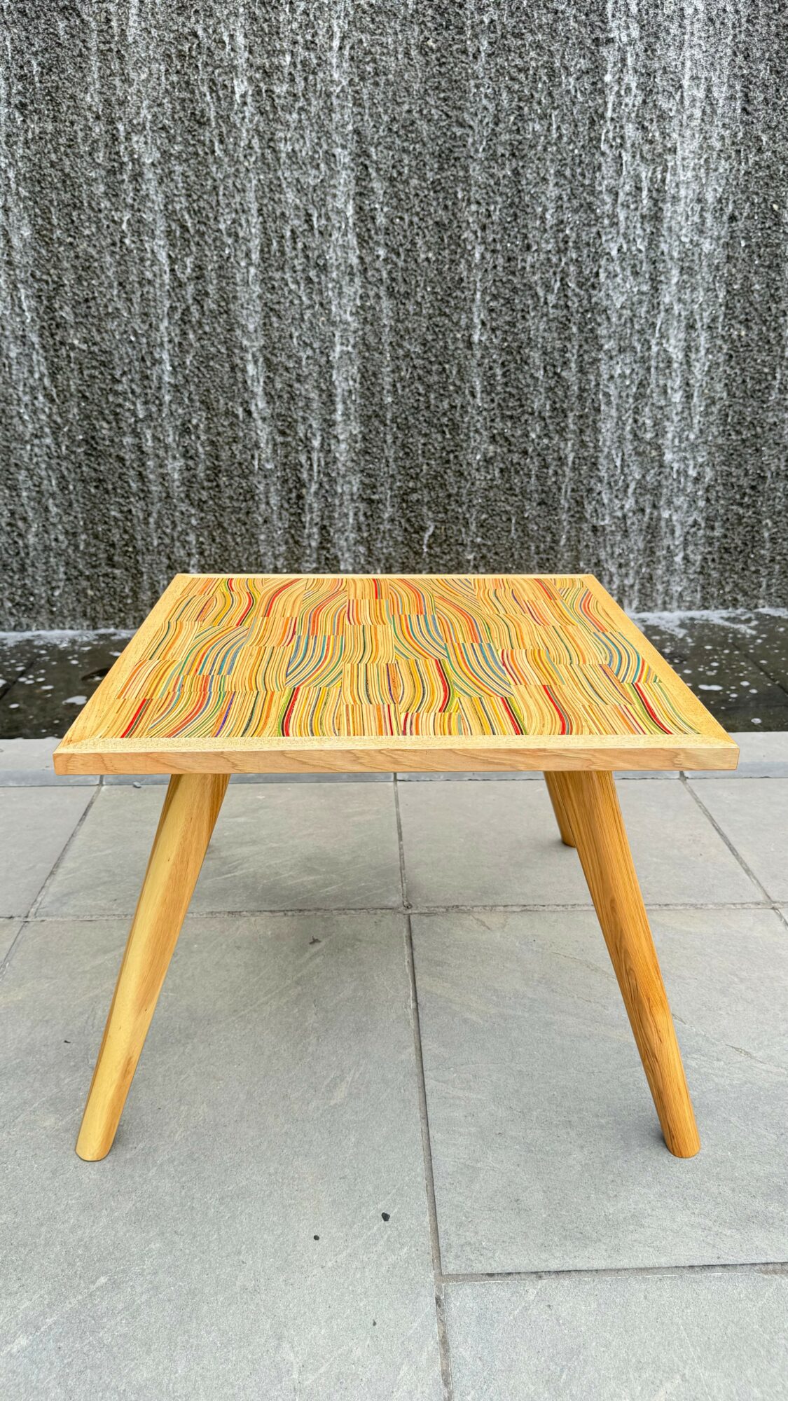 Small wooden table with colorful wood grain pattern, three angled legs, placed outdoors against a dark textured wall.
