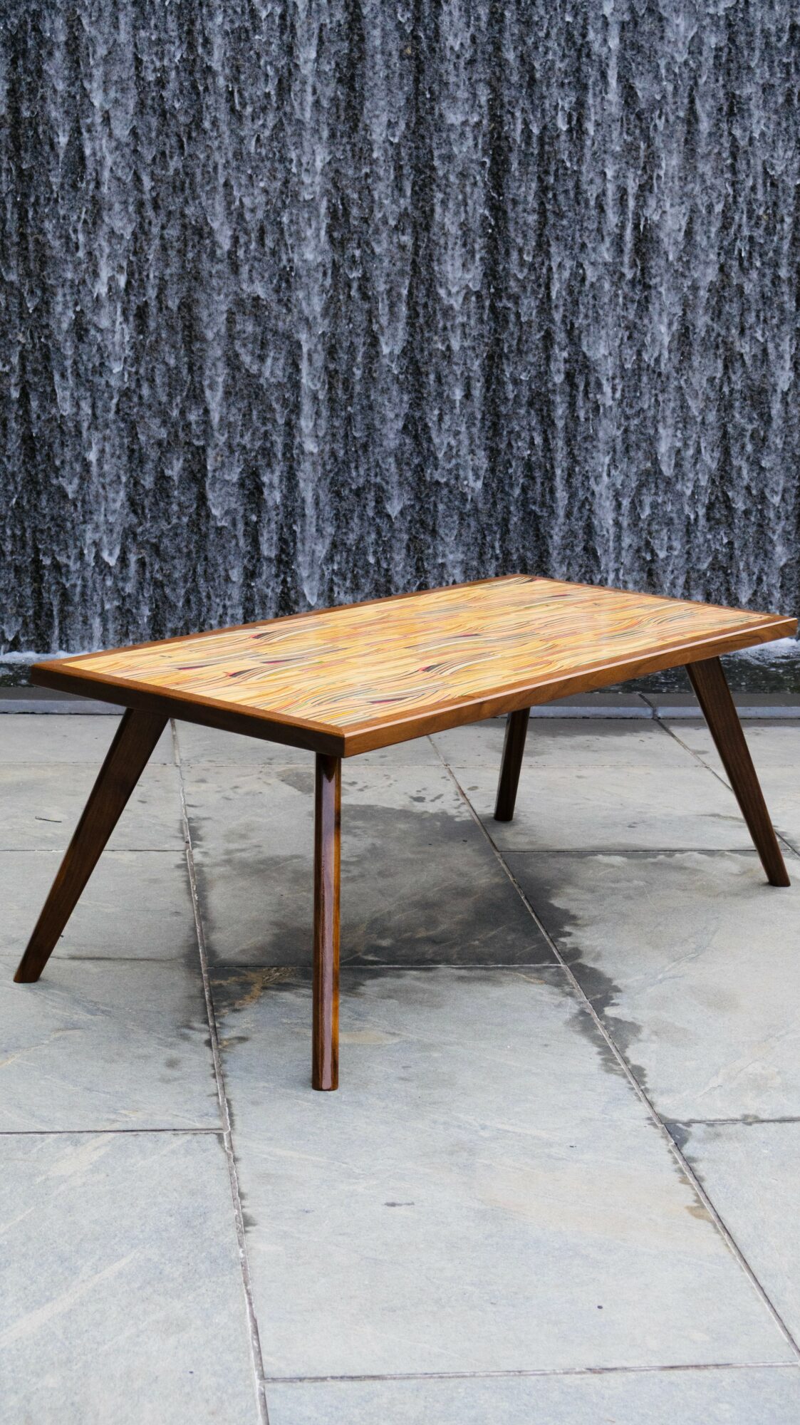 Wooden table with angled legs on a concrete surface, with a dark textured wall in the background.