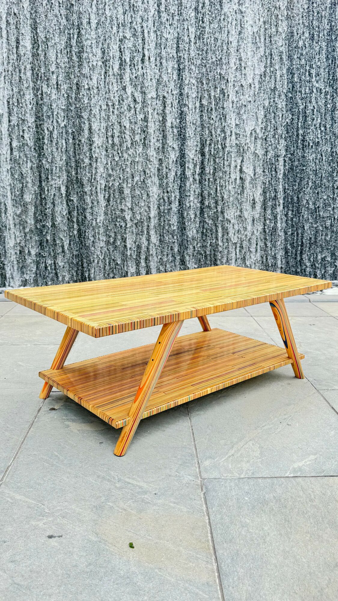Wooden table with a lower shelf, placed outdoors on a tiled surface, against a textured gray wall.